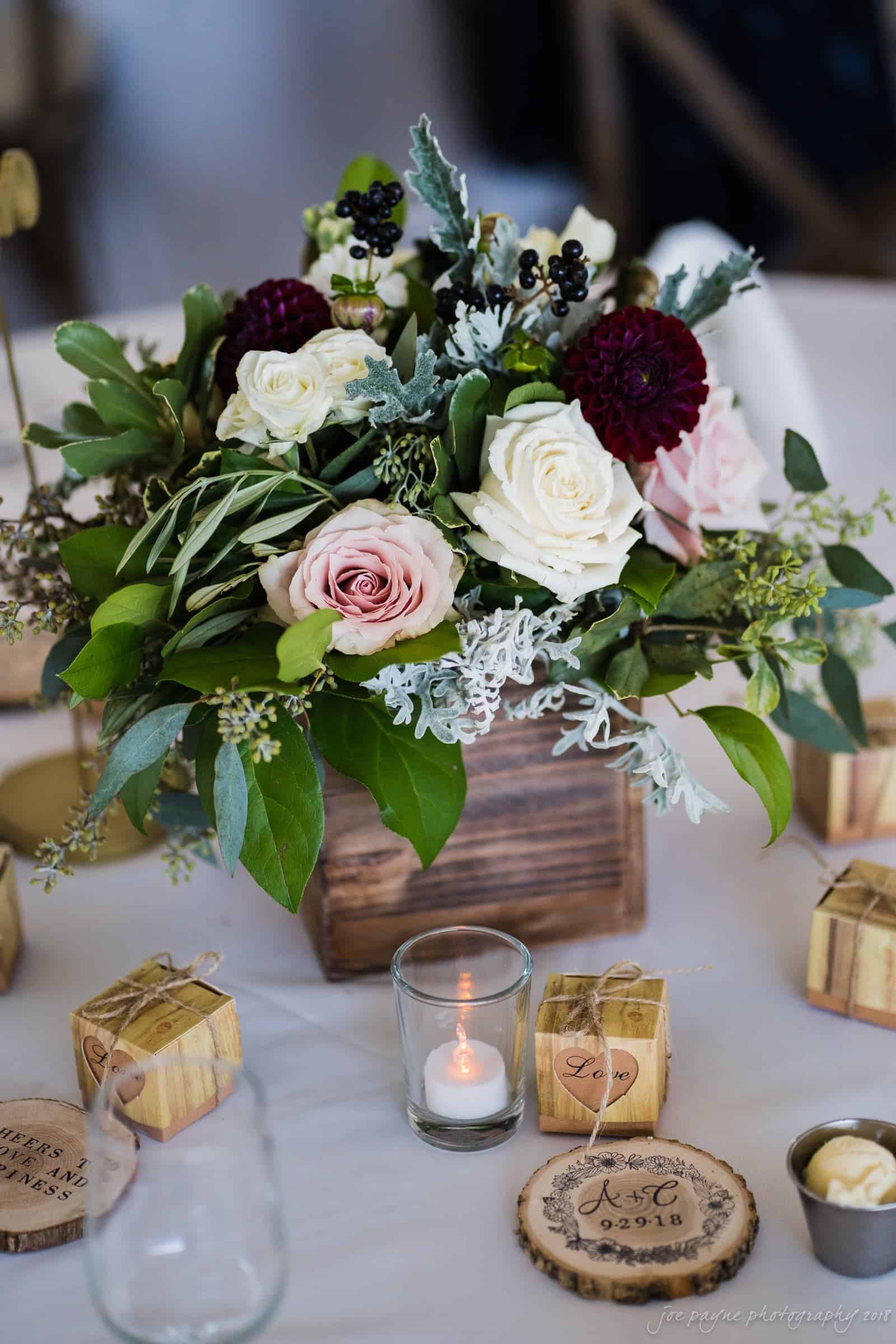 A rustic wooden box centerpiece holds a bouquet of white, pink, and dark purple roses with greenery. Surrounding it are small, wooden blocks with engraved details and tealight candles, arranged on a white tablecloth.