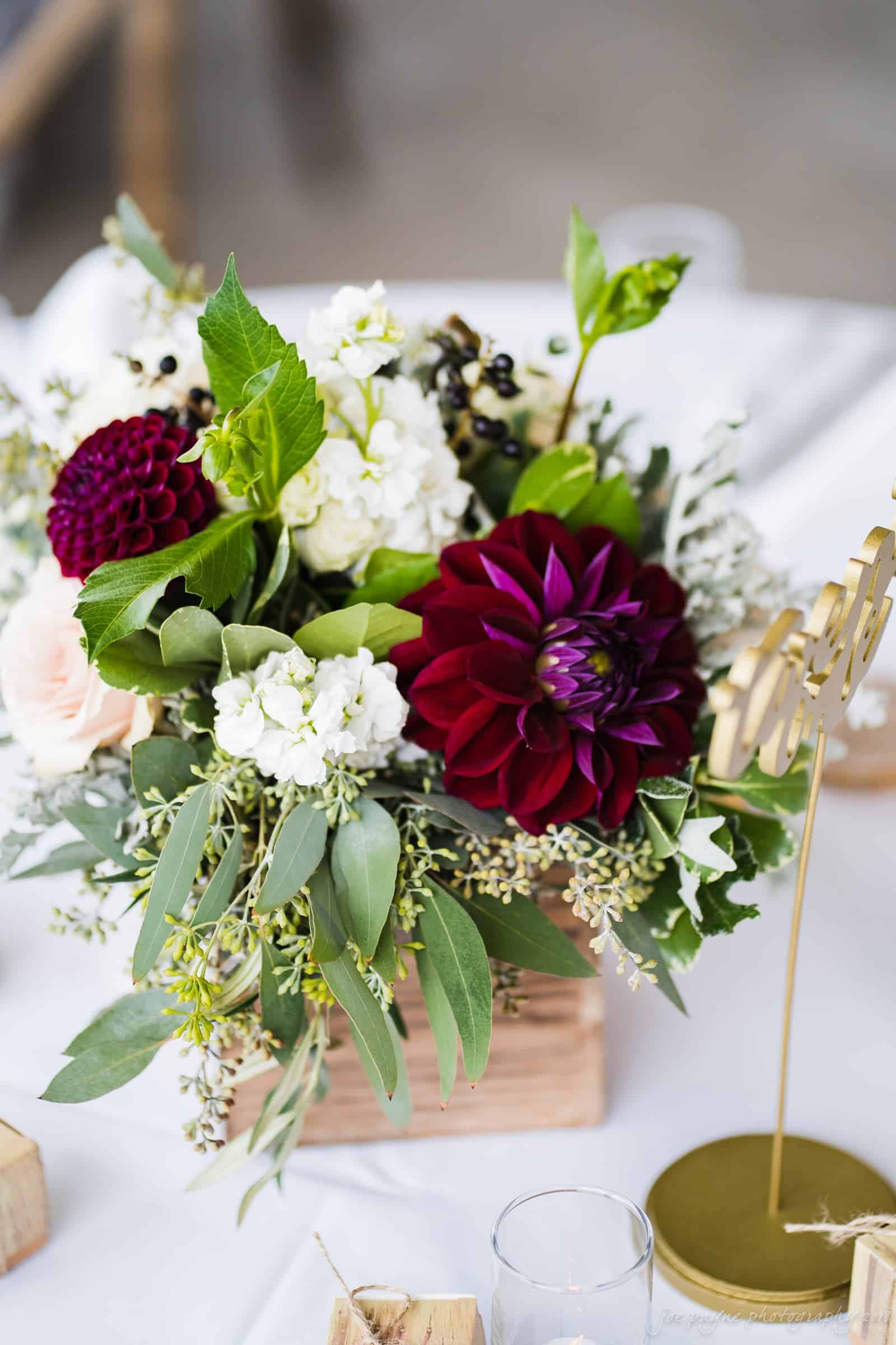 Close-up of a floral arrangement in a wooden box, featuring deep red dahlias, white flowers, leafy greens, and small berries. The arrangement is set on a white tablecloth with a gold decorative sign in the background.