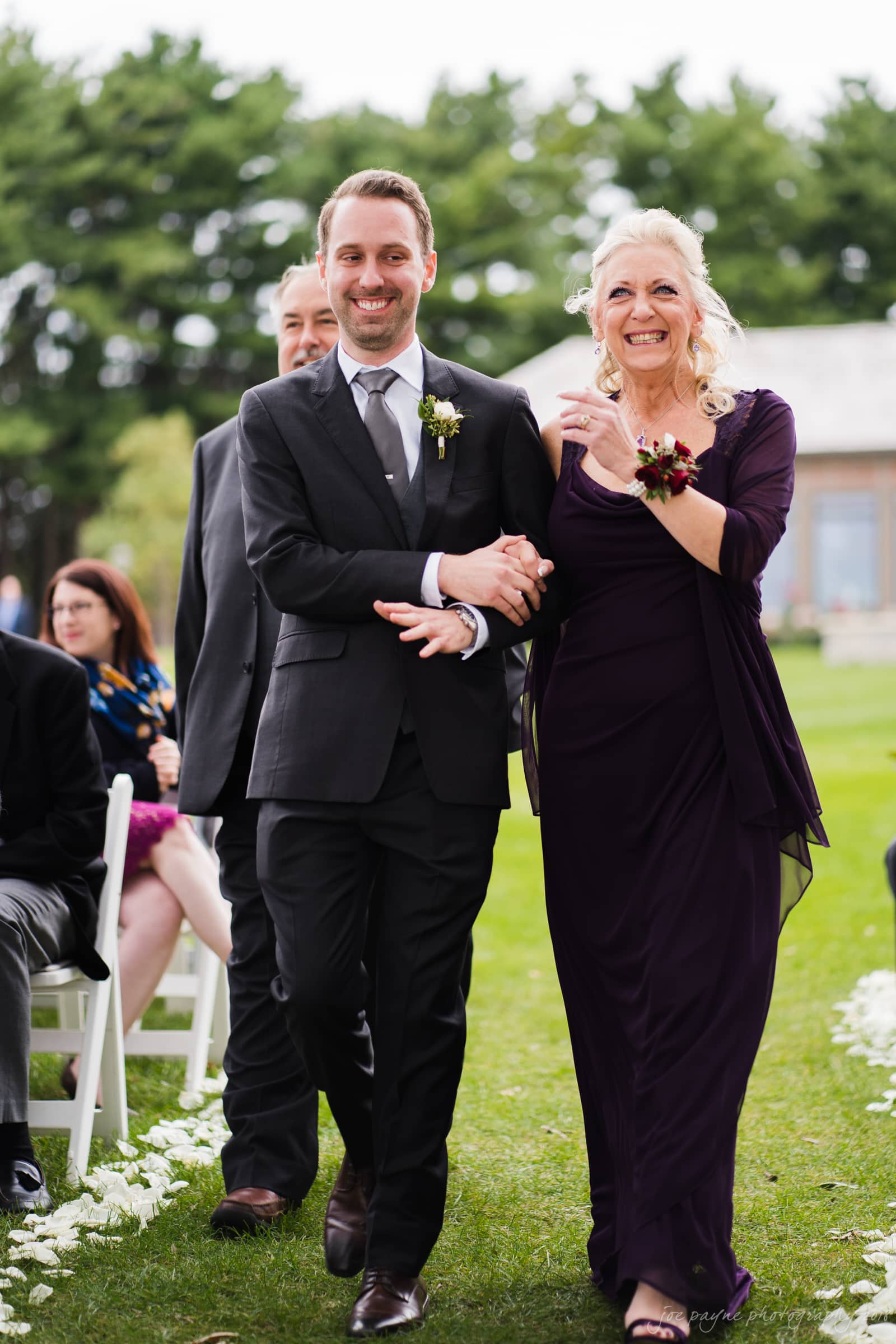 A smiling man in a suit walks arm-in-arm with a happy older woman in a dark dress and corsage. They are on a grassy path with seated guests and trees in the background, possibly at a wedding ceremony.