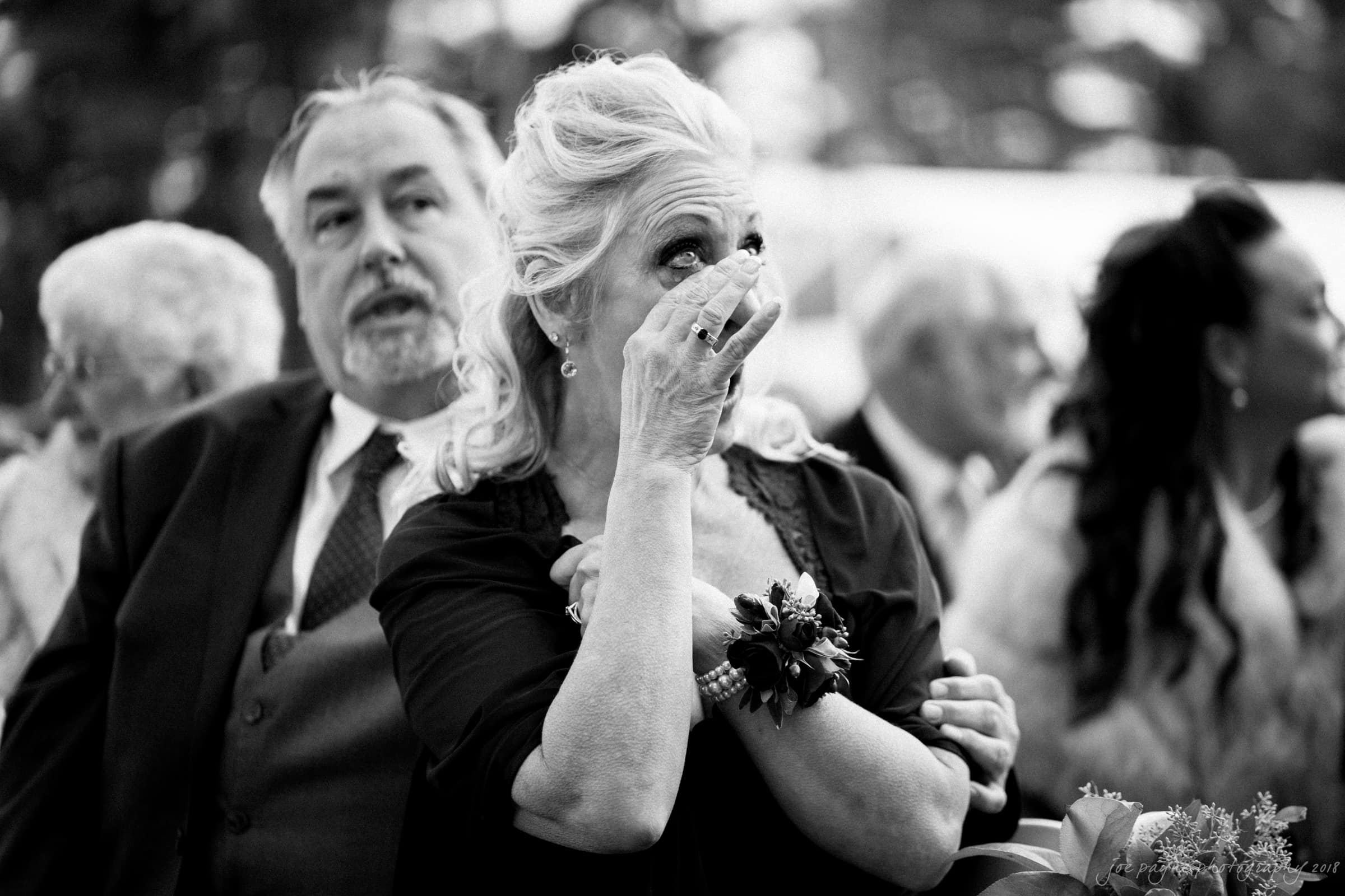 A black and white photo of an elderly woman wiping away a tear, wearing a floral corsage. She is surrounded by people, with a man standing behind her. The mood appears emotional, suggesting a sentimental moment at an event.