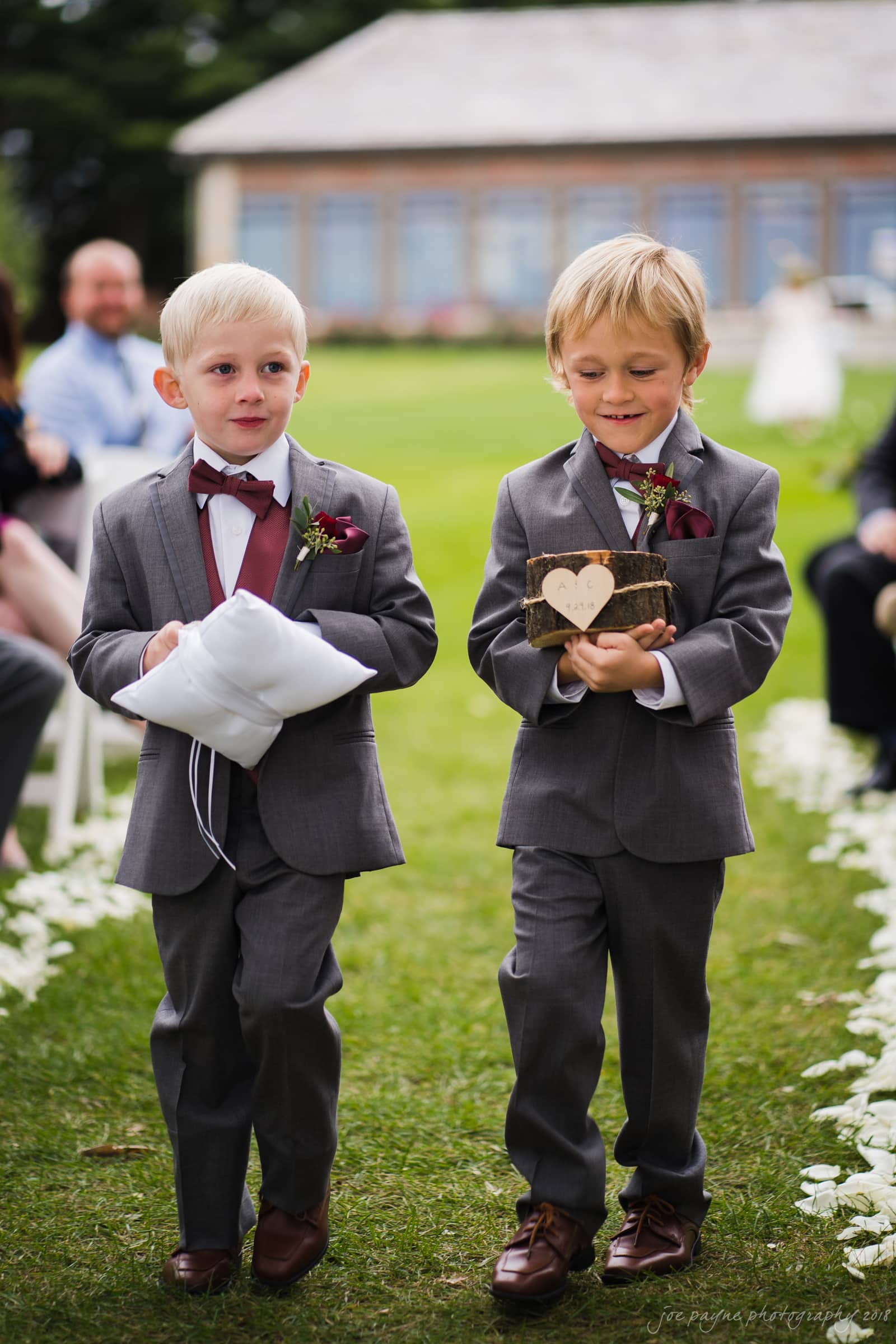 Two young boys in gray suits walk down an outdoor aisle. One holds a ring pillow, and the other carries a box with a heart decoration. Flower petals line the grass, and blurred guests are seated in the background.
