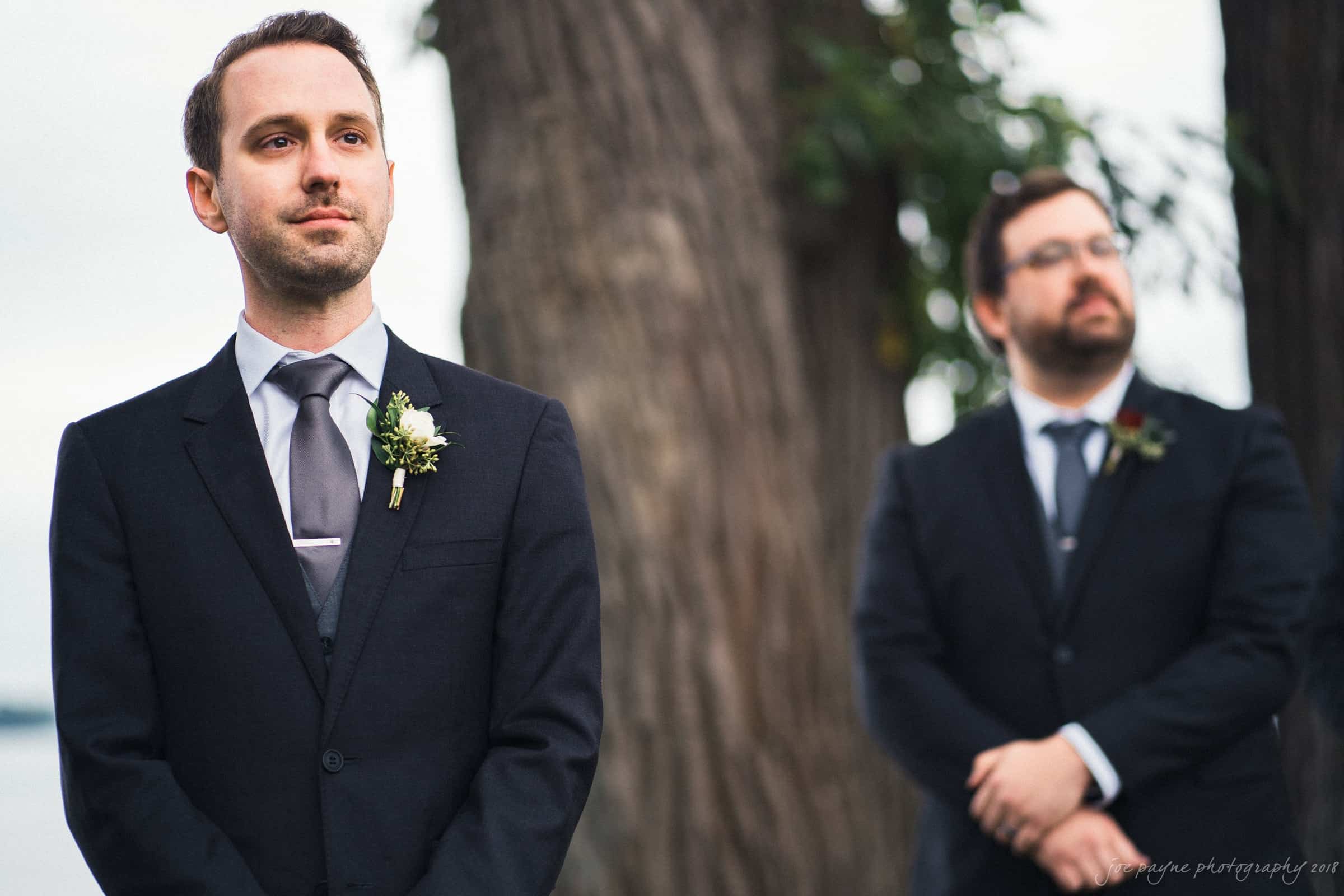 A groom stands outside in front of a large tree, wearing a dark suit, light shirt, and tie, with a white boutonniere. A man in a similar outfit is in the background, slightly blurred.