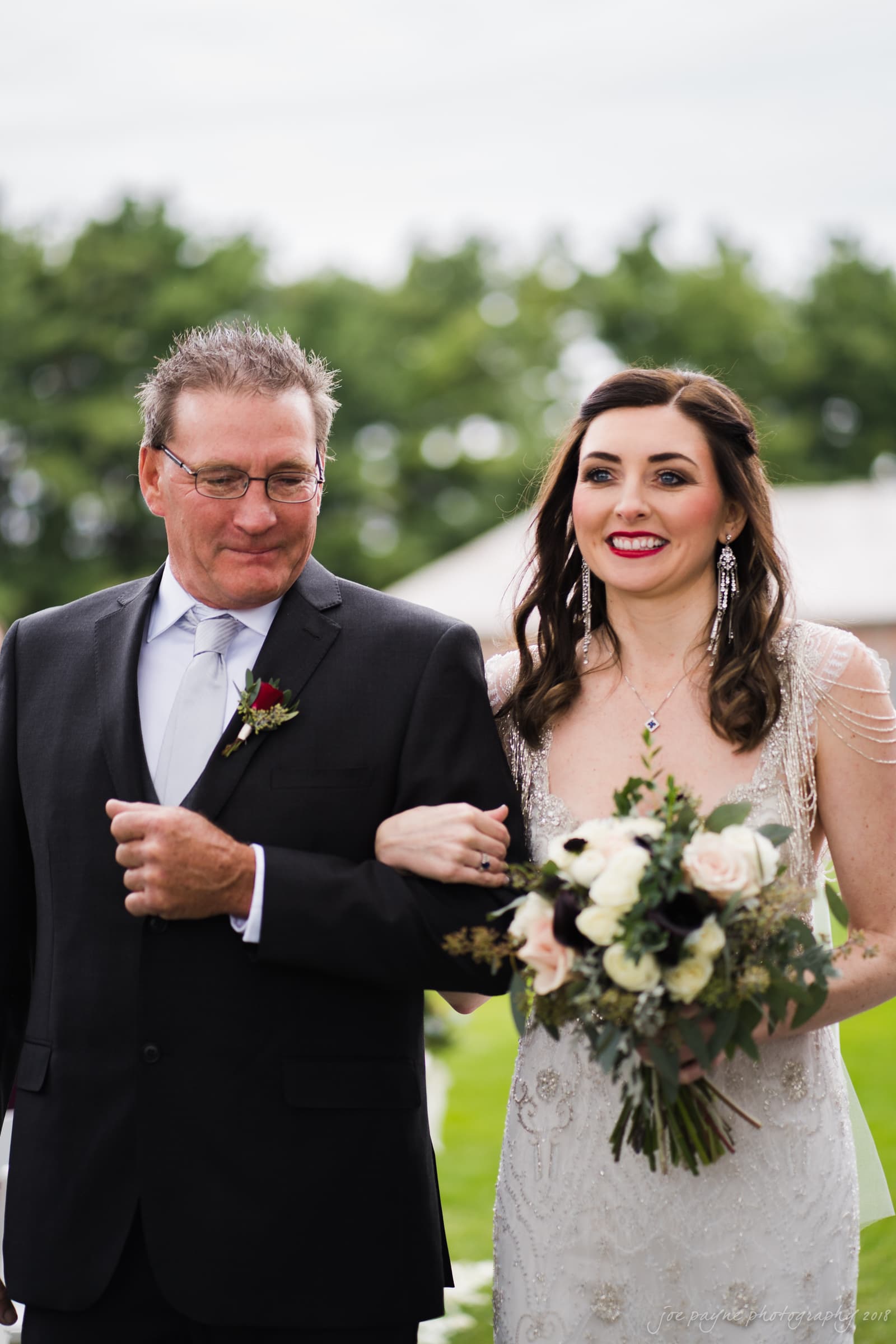 A bride in a white gown holds a bouquet of flowers as she walks arm in arm with an older man in a black suit and tie. They are outdoors, with greenery visible in the background. Both are smiling.