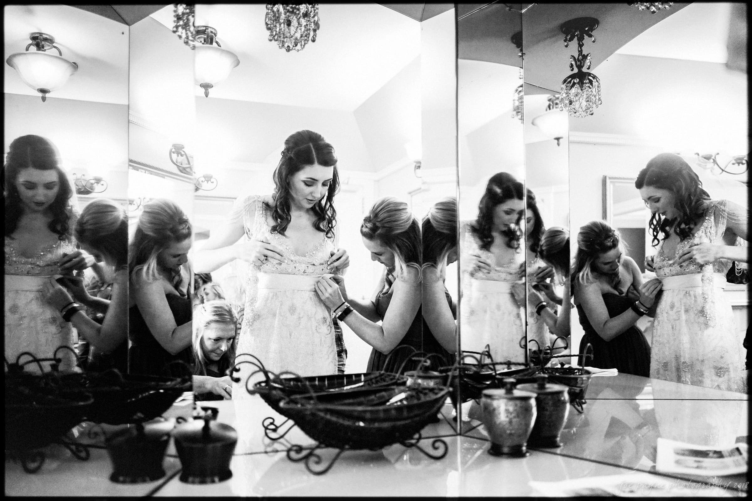Black and white image of a woman in a wedding dress being assisted by another woman. They are surrounded by mirrors, creating multiple reflections. The room is adorned with decorative light fixtures and bowls.