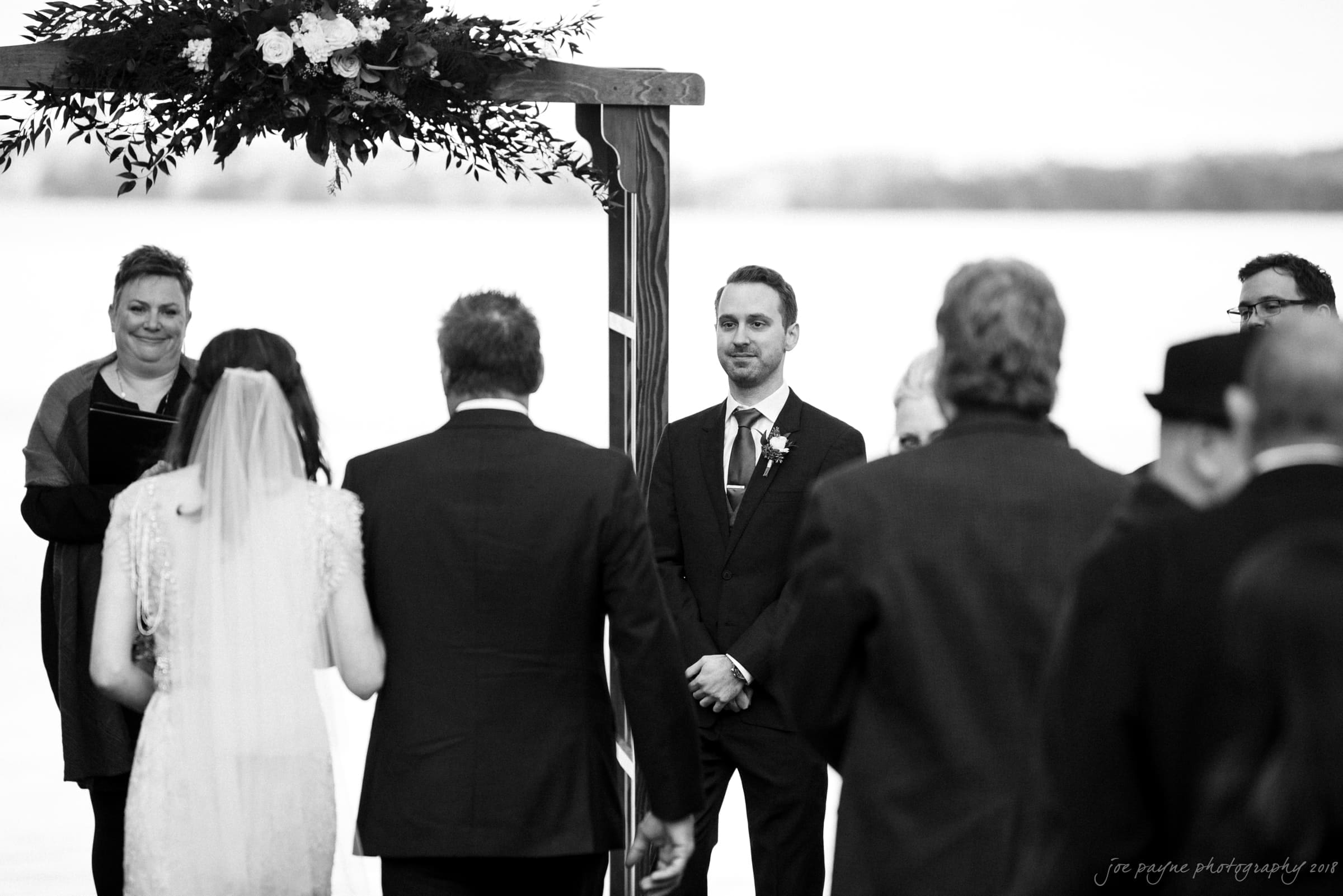A black and white photo of a wedding ceremony. The groom stands at the altar, smiling, while the bride approaches with her father. A floral arrangement decorates the altar, and an officiant stands to the side. Guests observe the scene.