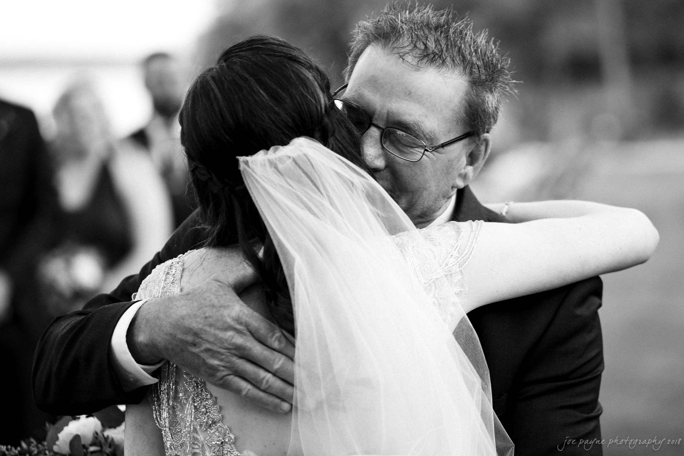 A joyful black and white image of a bride with a veil embracing an older man in a suit, both smiling warmly. The scene appears to be outdoors, with blurred guests in the background.