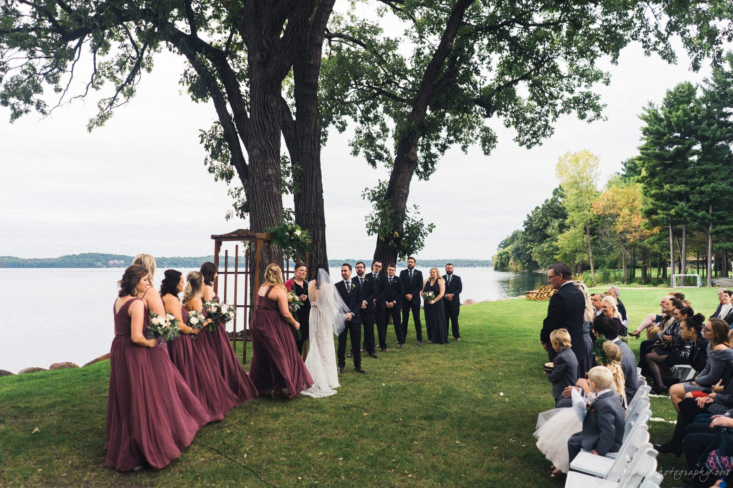 A wedding ceremony by a lake with trees and guests seated. The bride and groom stand under a wooden arch with bridesmaids in maroon dresses on the left and groomsmen in black suits on the right. The sky is overcast, and the setting is lush with greenery.