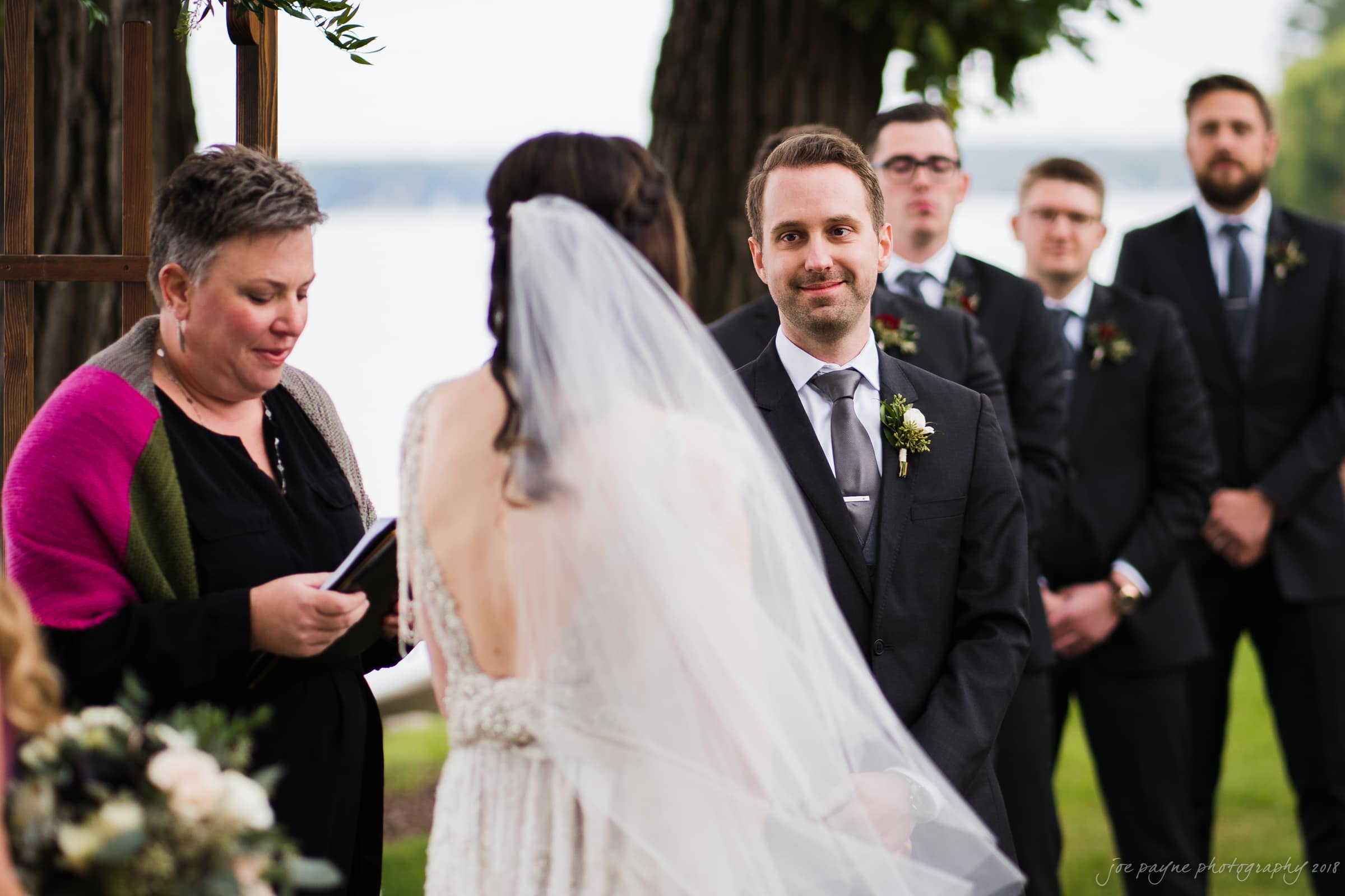 A wedding ceremony by a lake, featuring a couple exchanging vows. The bride is in a white dress with a long veil, and the groom is in a dark suit with a boutonniere. An officiant and groomsmen stand by, with greenery and water in the background.