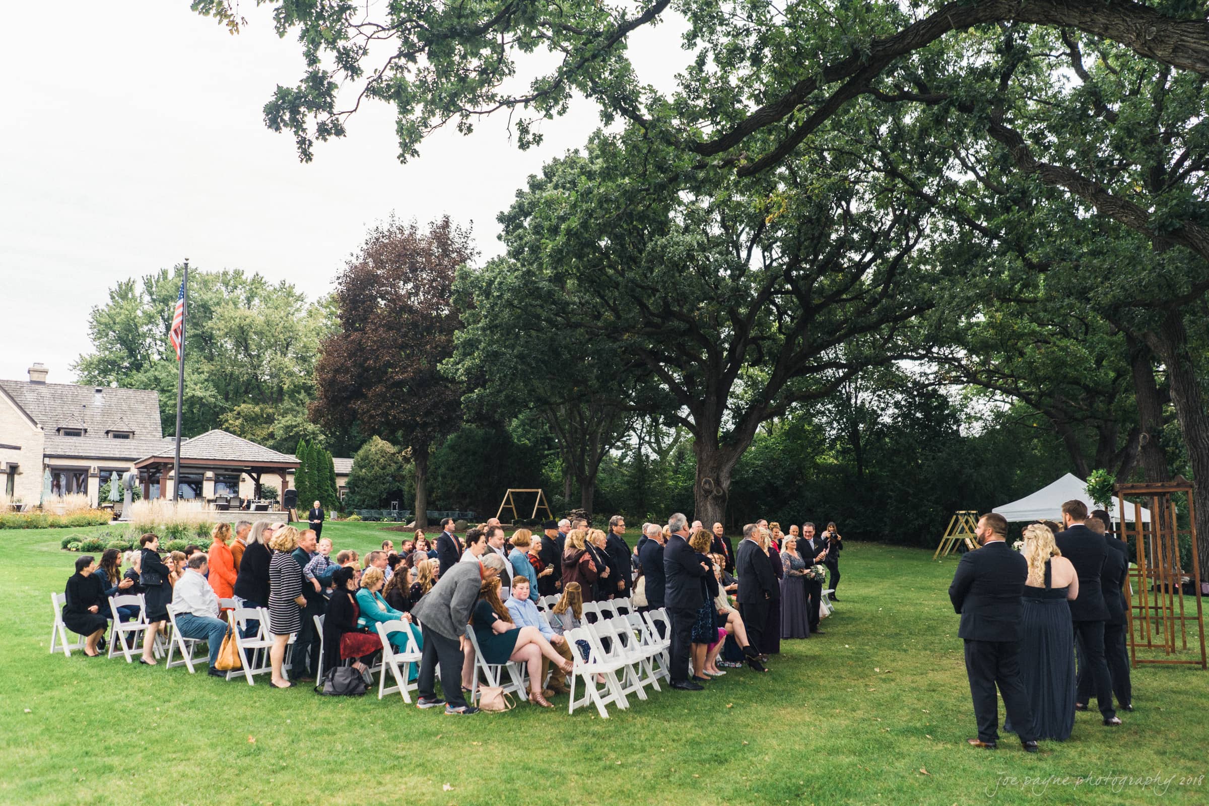 A wedding ceremony is taking place outdoors under tall trees. Guests are seated on white folding chairs, facing an altar. The setting is a green, grassy lawn, with a large house and flagpole visible in the background.