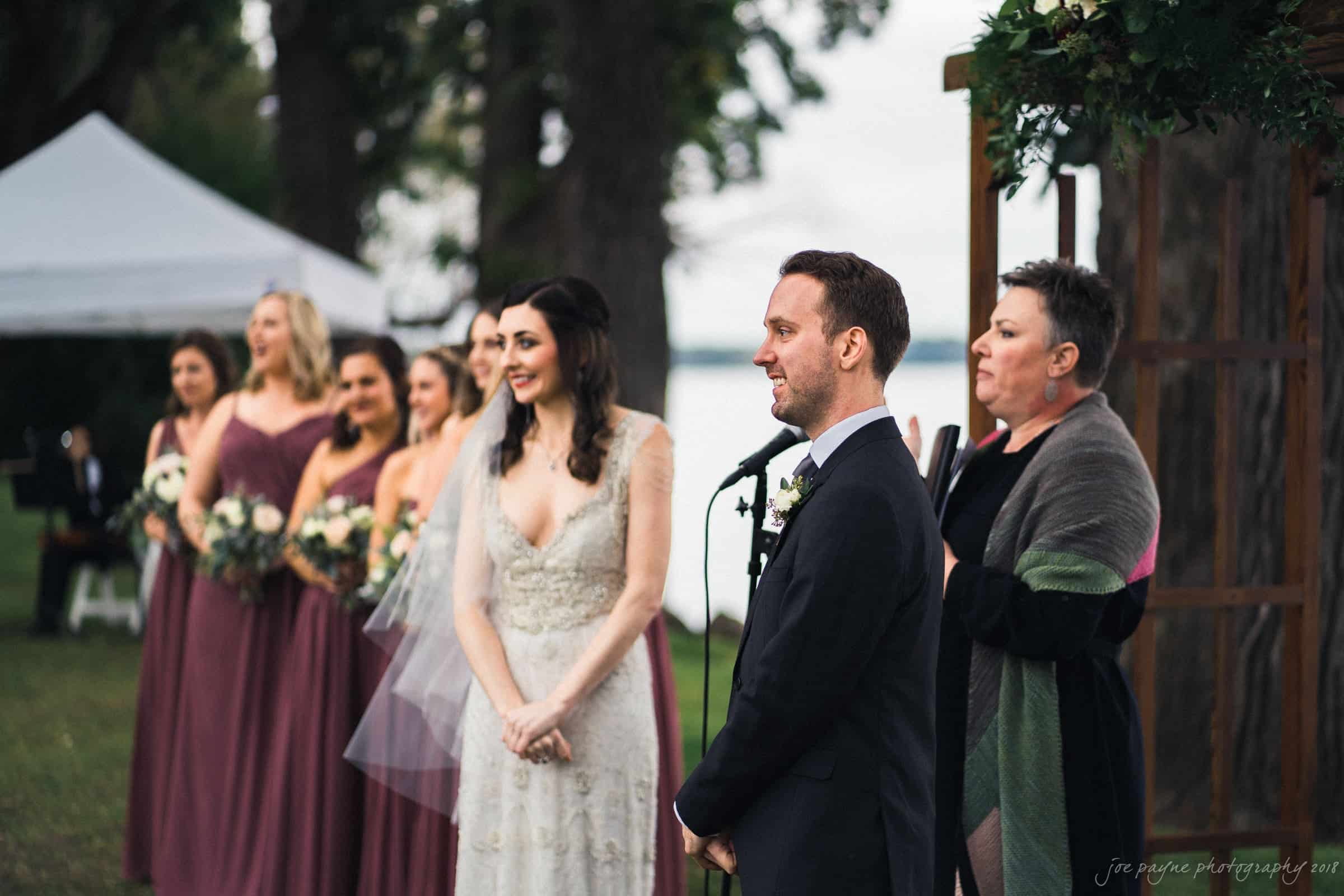 A bride and groom stand outdoors at a wedding ceremony. The bride wears a lacy gown and veil, while the groom is in a suit with a boutonniere. The officiant stands beside them. Bridesmaids in matching dresses hold bouquets in the background.