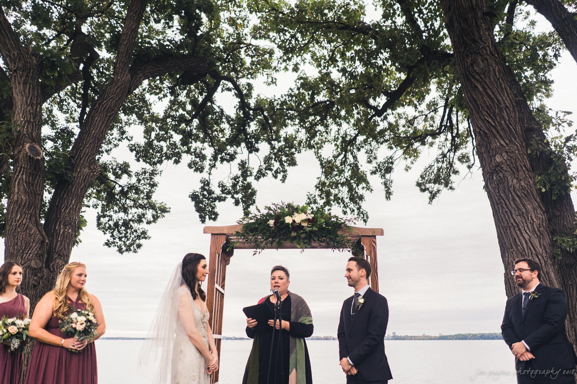 A wedding ceremony by a lake, with a bride in a white dress and veil and a groom in a suit standing under a wooden arch decorated with greenery. The officiant stands between them, surrounded by four attendants. Large trees frame the scene.
