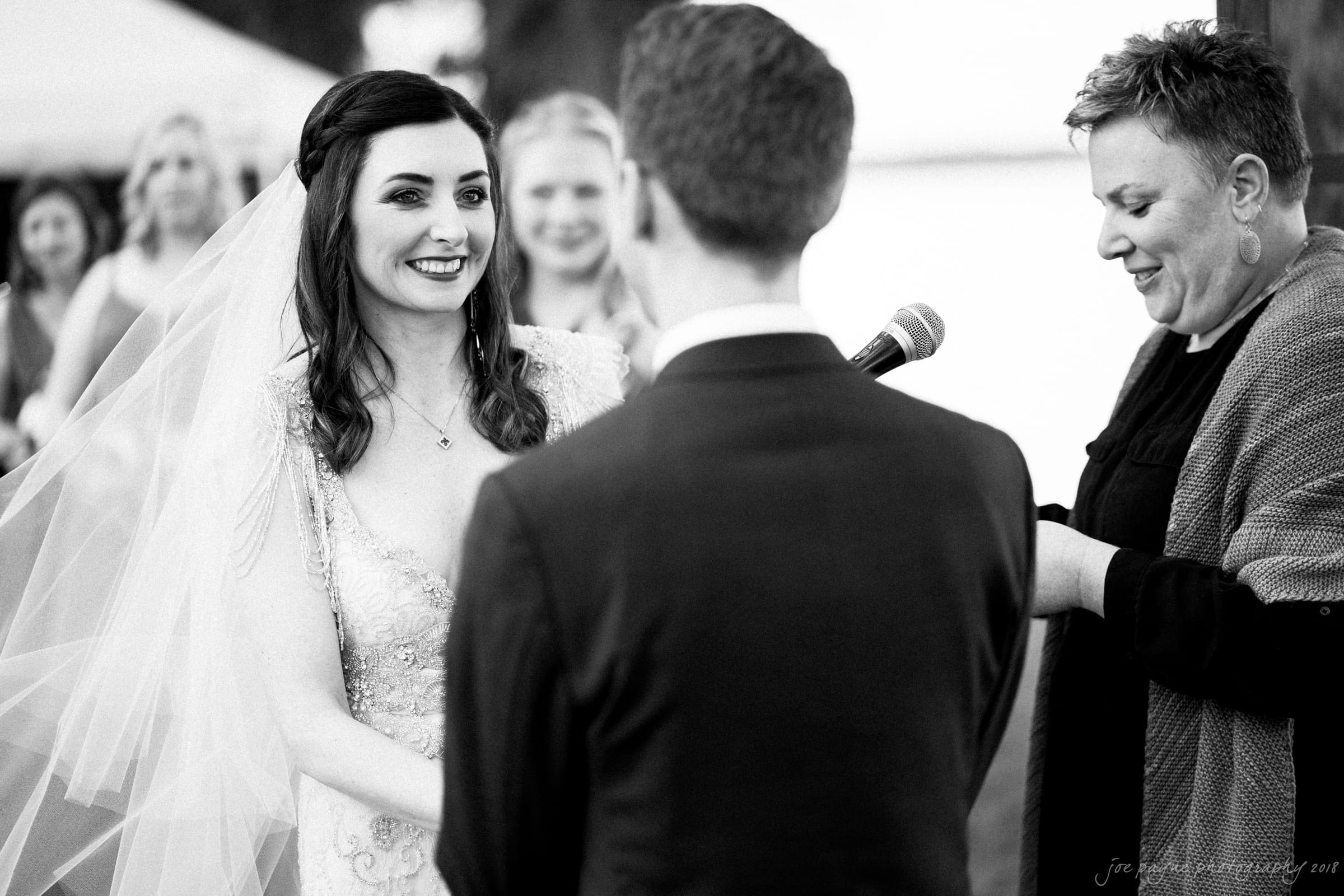 A bride and groom stand facing each other during an outdoor wedding ceremony. The bride is smiling and wearing a veil, while a woman speaks into a microphone. Other people are visible in the background. Black and white photo.
