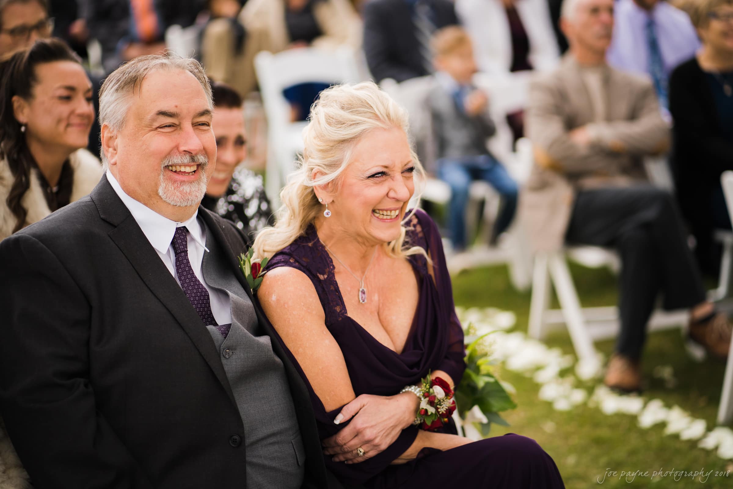 A joyful couple sitting together, dressed elegantly for a special occasion. The man wears a dark suit, and the woman is in a purple dress, both smiling and surrounded by seated guests outdoors. White chairs and blurred attendees are visible in the background.