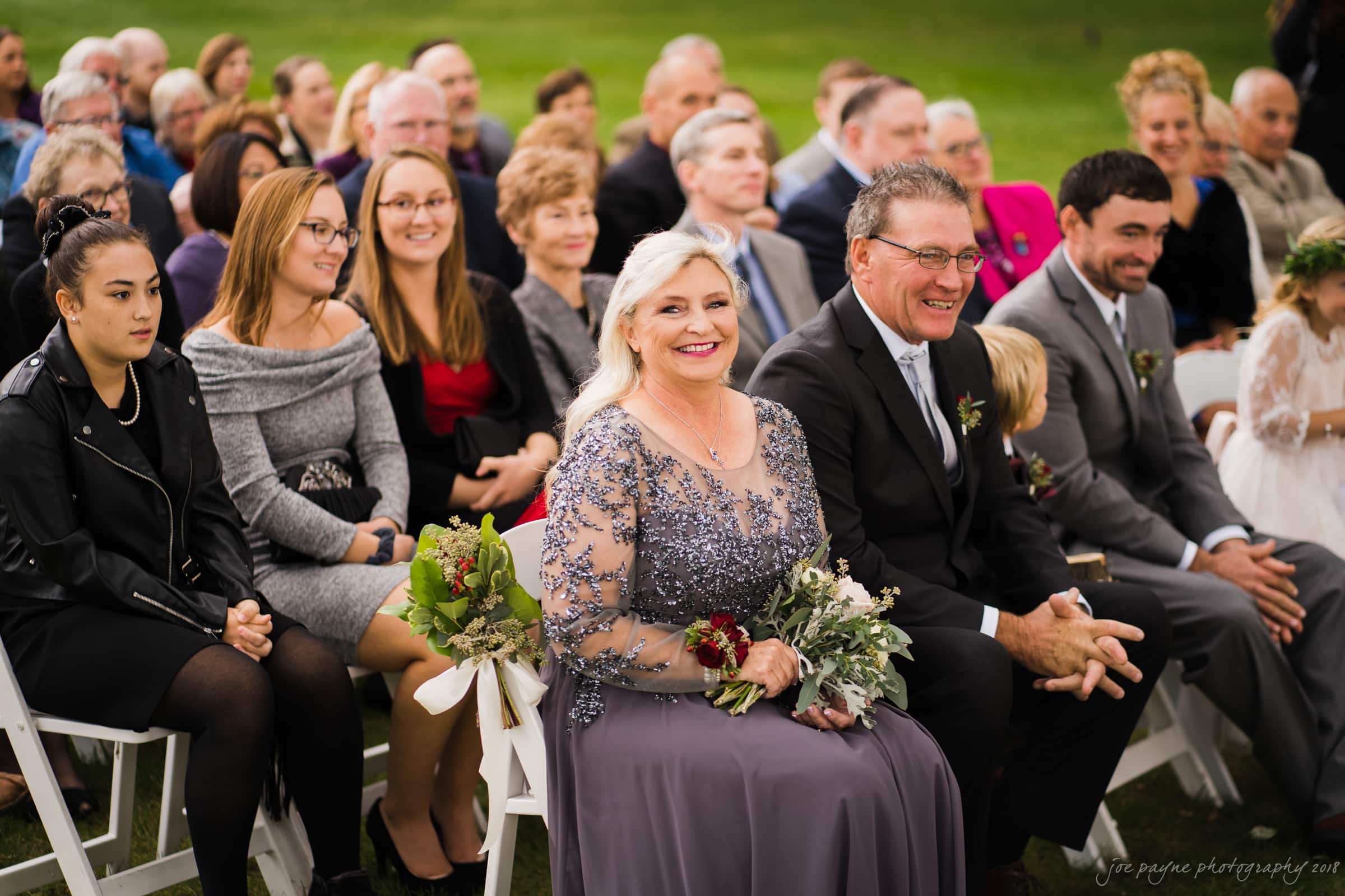 A group of smiling guests seated in rows at an outdoor event. In the foreground, two older individuals hold bouquets. Everyone appears happy and engaged, with a grassy background suggesting a ceremony or celebration.