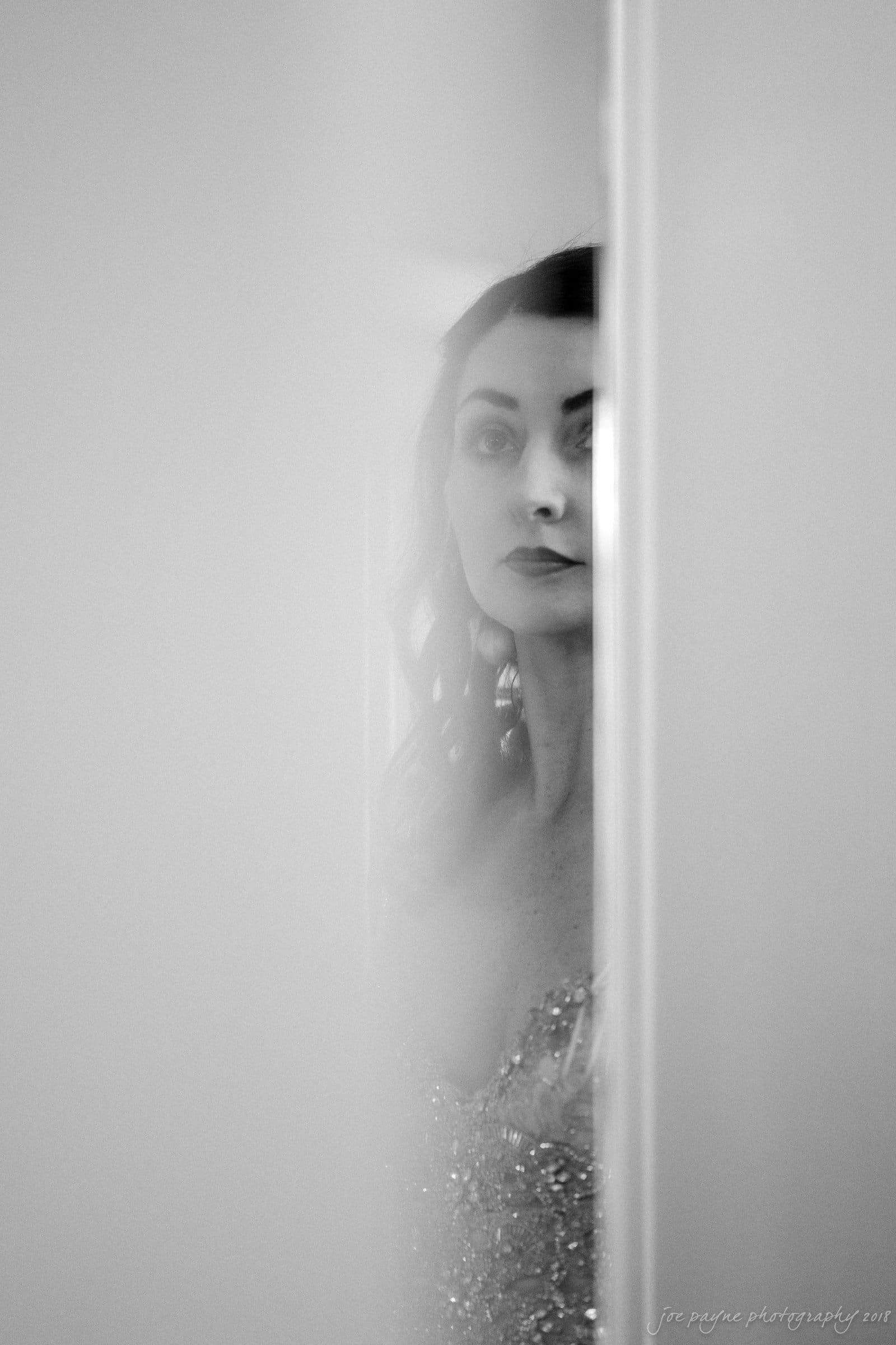 A black-and-white photo shows a woman partially obscured by a wall. She has long hair and wears a sparkly top, gazing forward with a soft expression. The image is artistically blurred for a dreamy effect.