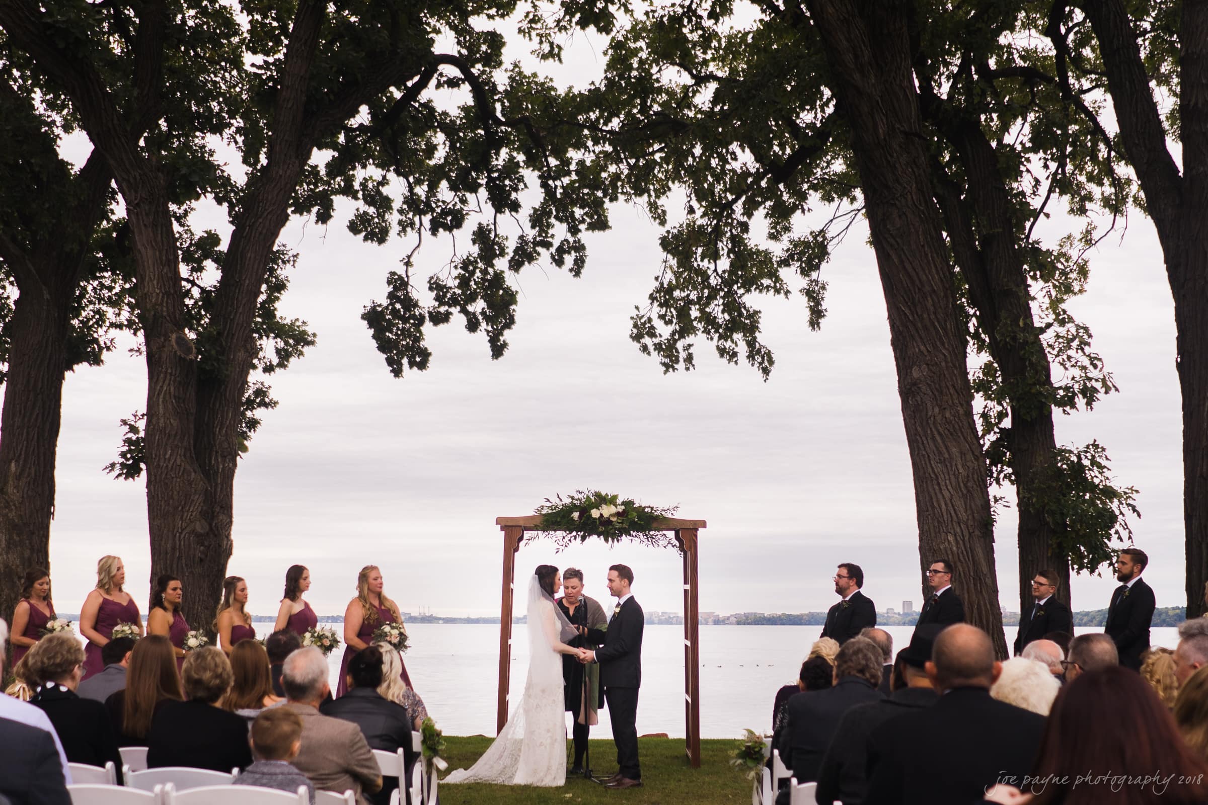 A wedding ceremony outdoors by a lake, with a couple standing under a wooden arch adorned with greenery. Guests are seated on either side, and the wedding party stands nearby, surrounded by tall trees.