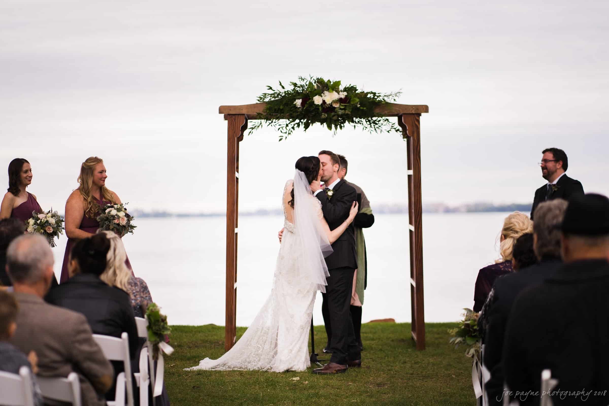 A couple kisses under an arbor at an outdoor wedding ceremony by a lake. The bride wears a white dress and the groom a dark suit. Bridesmaids in matching dresses and guests seated on white chairs watch the scene. The sky is overcast.