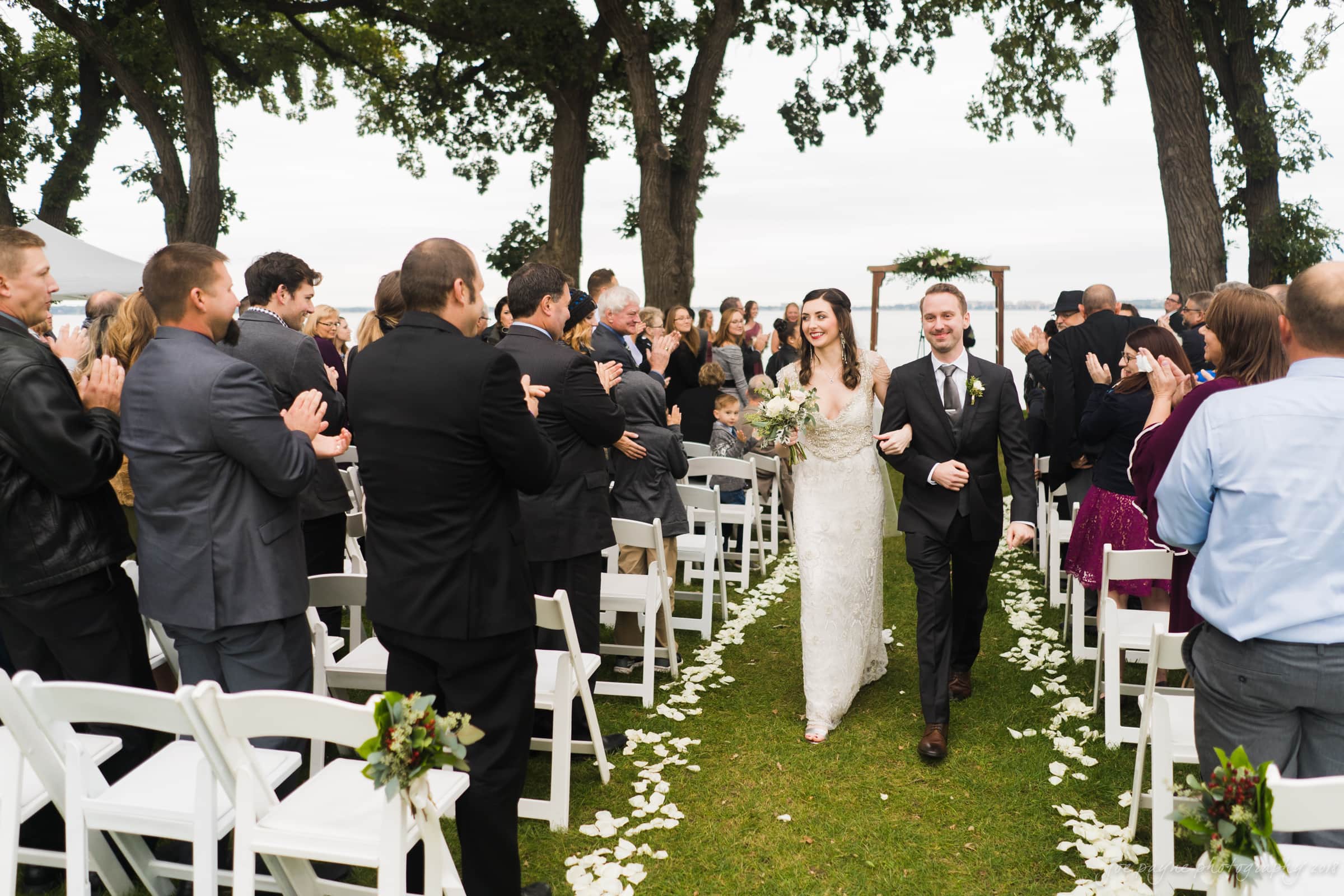 A newlywed couple joyfully walks down an outdoor aisle lined with white chairs. Guests stand and clap. A floral archway is visible in the background, with tall trees framing the scene. The bride holds a bouquet as they smile together.