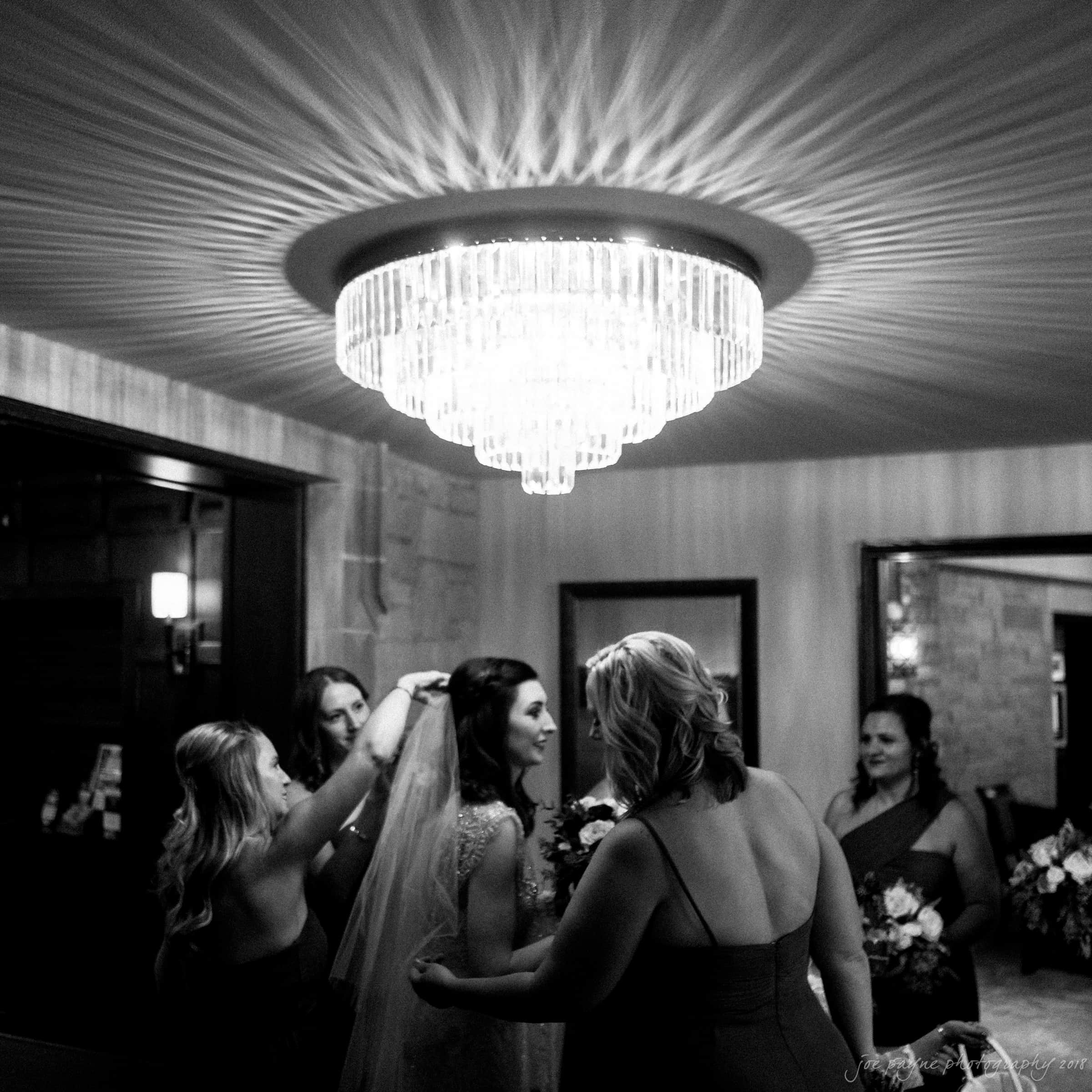 A bride stands under a sparkling chandelier while bridesmaids help with her veil. The room is elegantly lit, casting an ornate pattern on the ceiling. The bridesmaids hold bouquets, creating an atmosphere of celebration and anticipation.