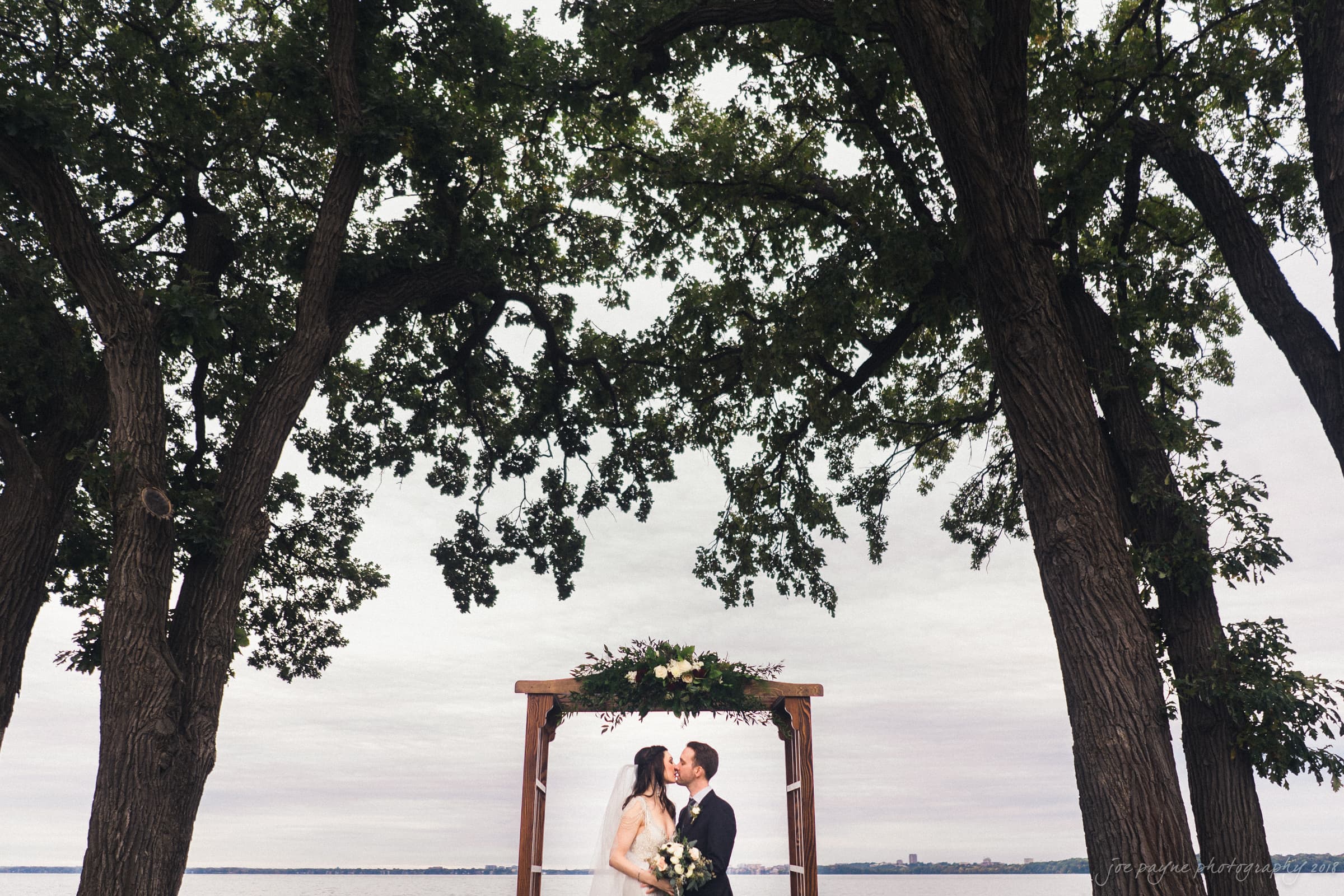 A couple kisses under a wooden arch adorned with flowers, surrounded by tall trees. The background features a cloudy sky and a distant view of water. The setting conveys a romantic outdoor atmosphere.