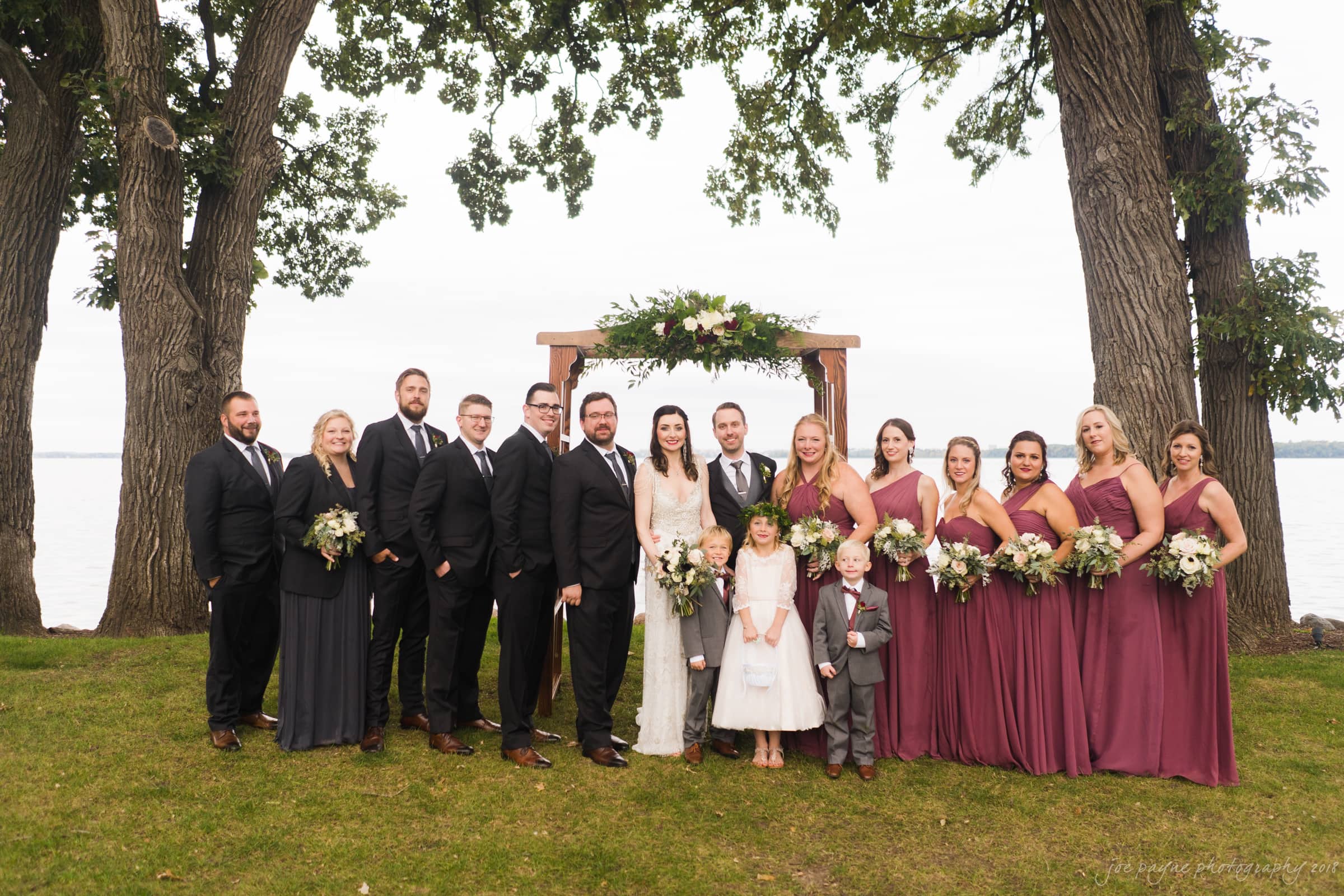 A wedding party stands outdoors by a wooden arbor adorned with flowers. The bride and groom are centered, surrounded by bridesmaids in burgundy dresses and groomsmen in black suits. Two children stand in front. Trees and a body of water are in the background.