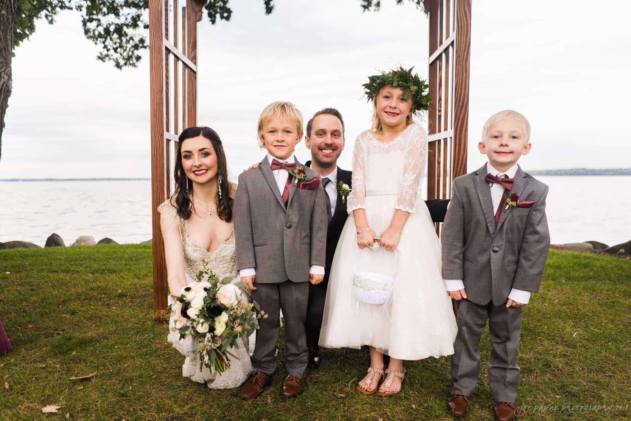 A bride and groom pose with three children in front of a wooden arch by a lakeside. The bride holds a bouquet, and the children, two boys in gray suits and a girl in a white dress with a floral crown, smile together on the grass.