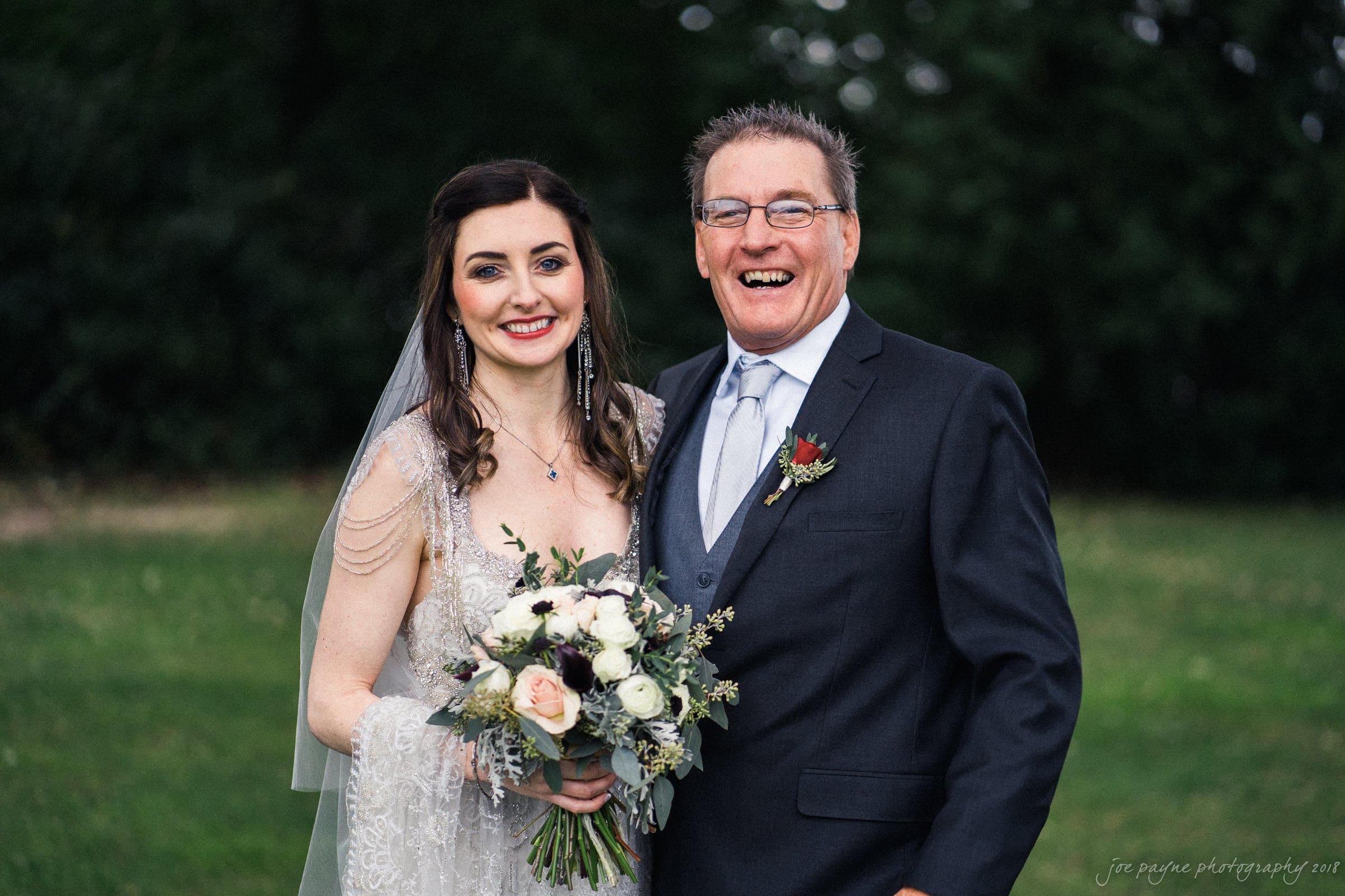 A bride in a white lace gown and veil stands smiling next to a man in a dark suit with a boutonniere. She holds a bouquet of white and pink flowers. They are outdoors on a grassy area with trees in the background.