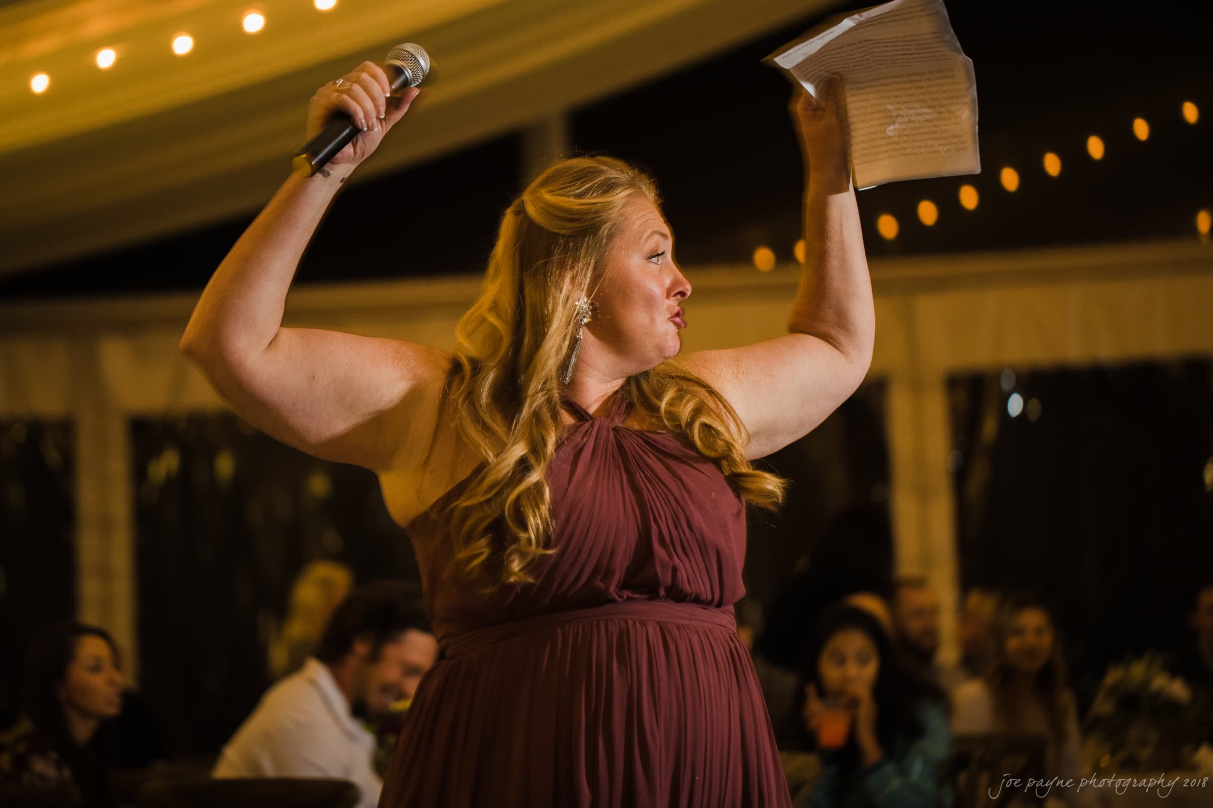 A woman in a sleeveless burgundy dress holds a microphone and a piece of paper, speaking passionately at an event. She gestures energetically, while seated guests in the dimly lit venue listen attentively. String lights hang across the ceiling.