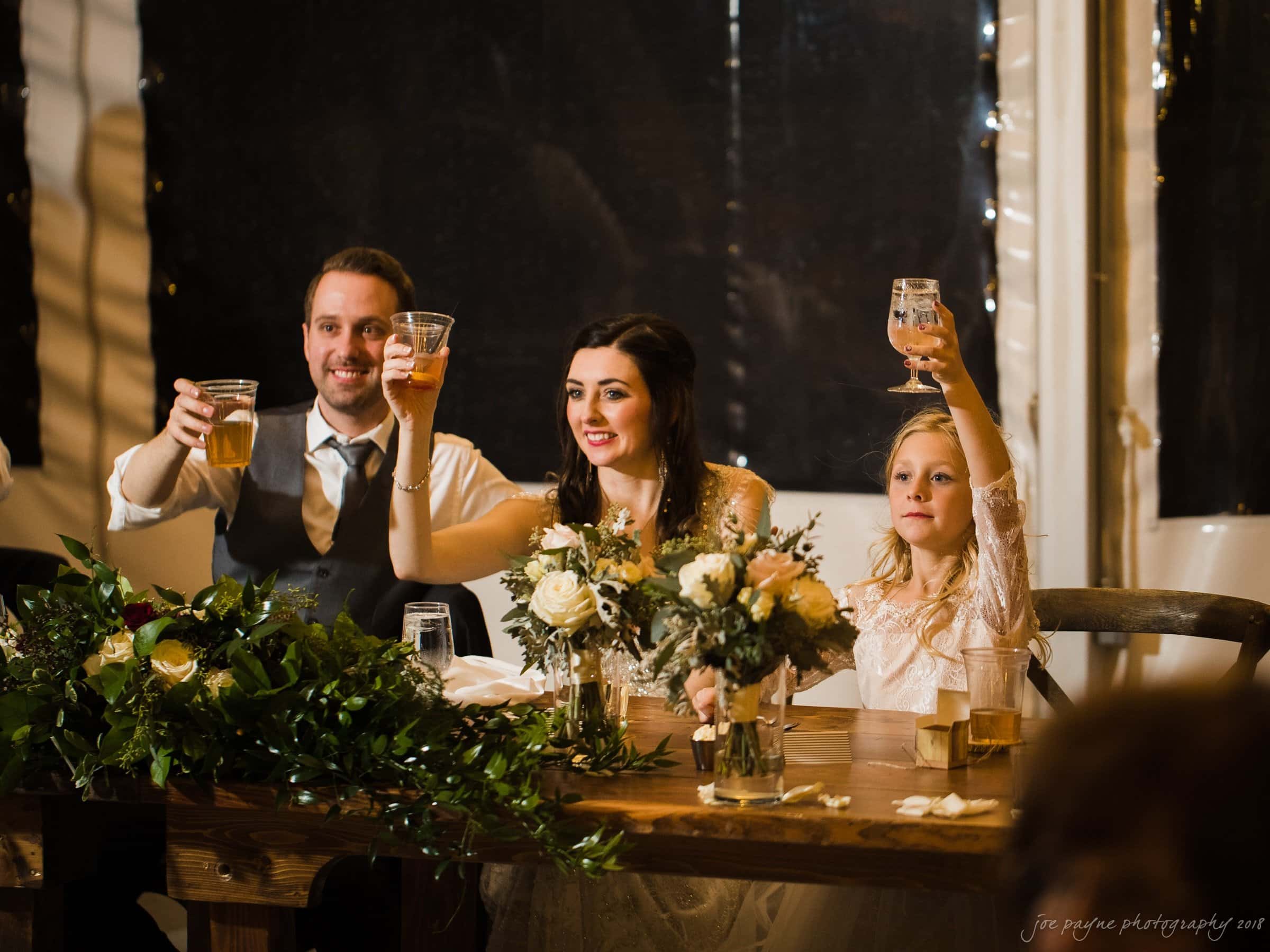 A bride, groom, and a young girl raise glasses in a celebratory toast at a wedding reception. They are seated at a wooden table adorned with floral arrangements. The atmosphere is warm and joyful.