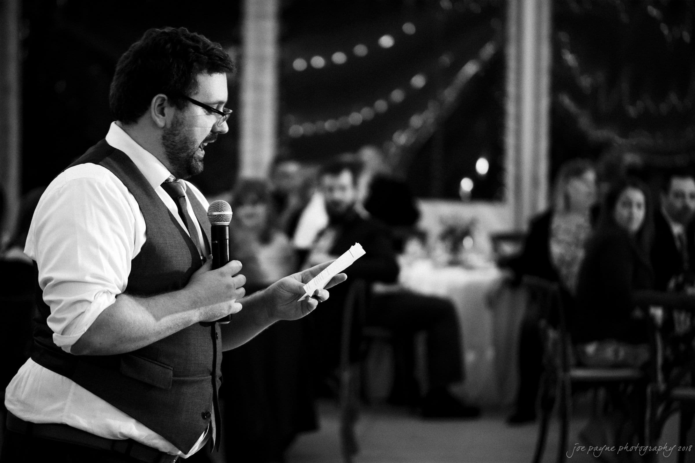 A man in a vest and tie speaks into a microphone while reading from a paper at a formal event. The background is softly blurred, showing seated guests at tables decorated with lights.