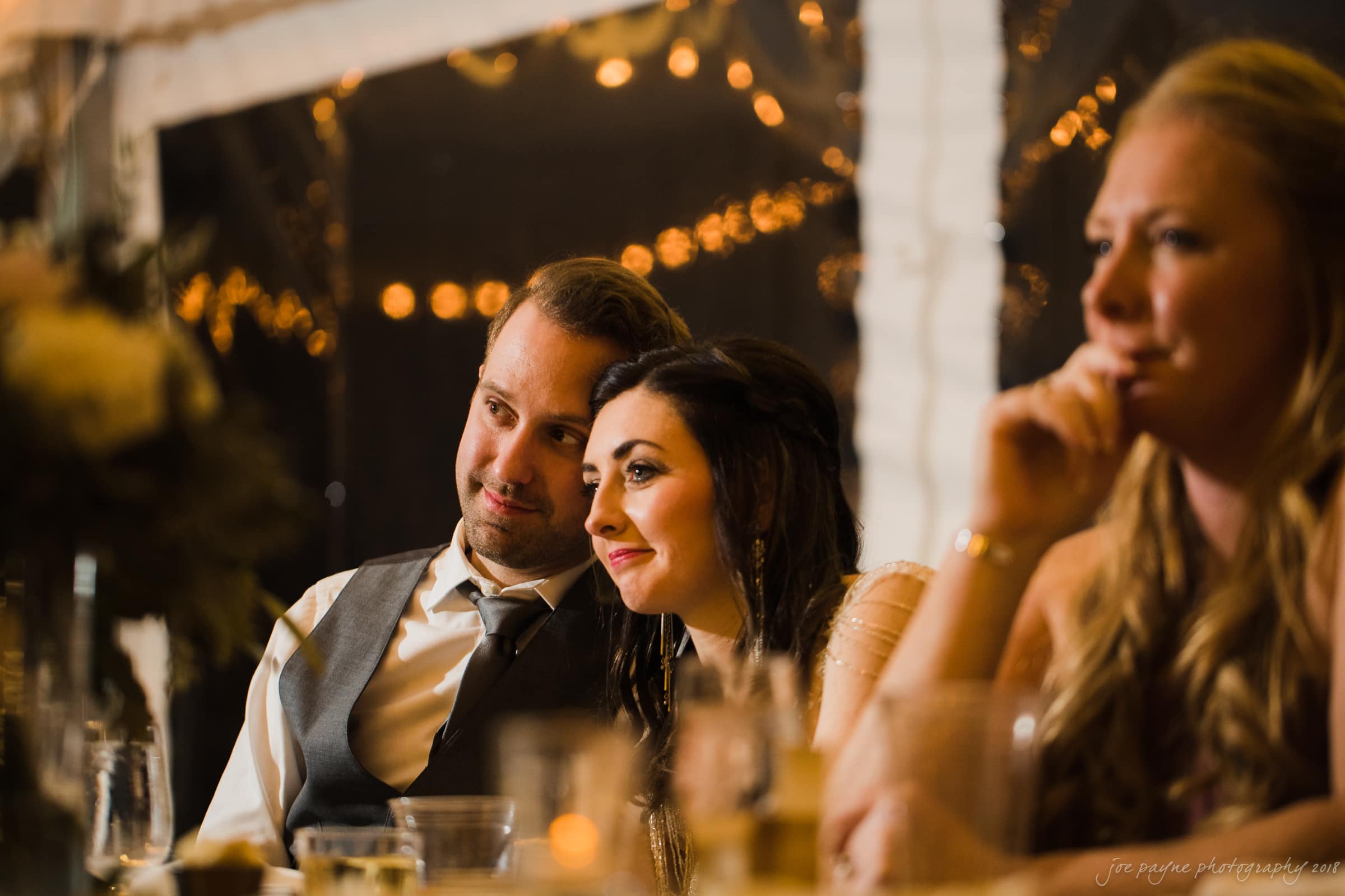 A couple sitting closely at a table during a warmly lit event. The man in a vest and tie leans towards the woman in a dress, who is smiling. Another person, a woman, sits to their right, partially visible. Glasses and decorations are on the table.