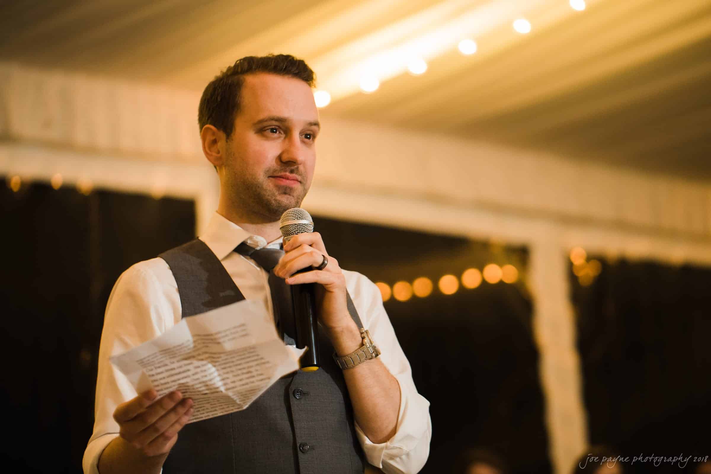 A man in a vest and tie is speaking into a microphone while holding a paper. He is standing indoors, with warm lighting and string lights in the background.