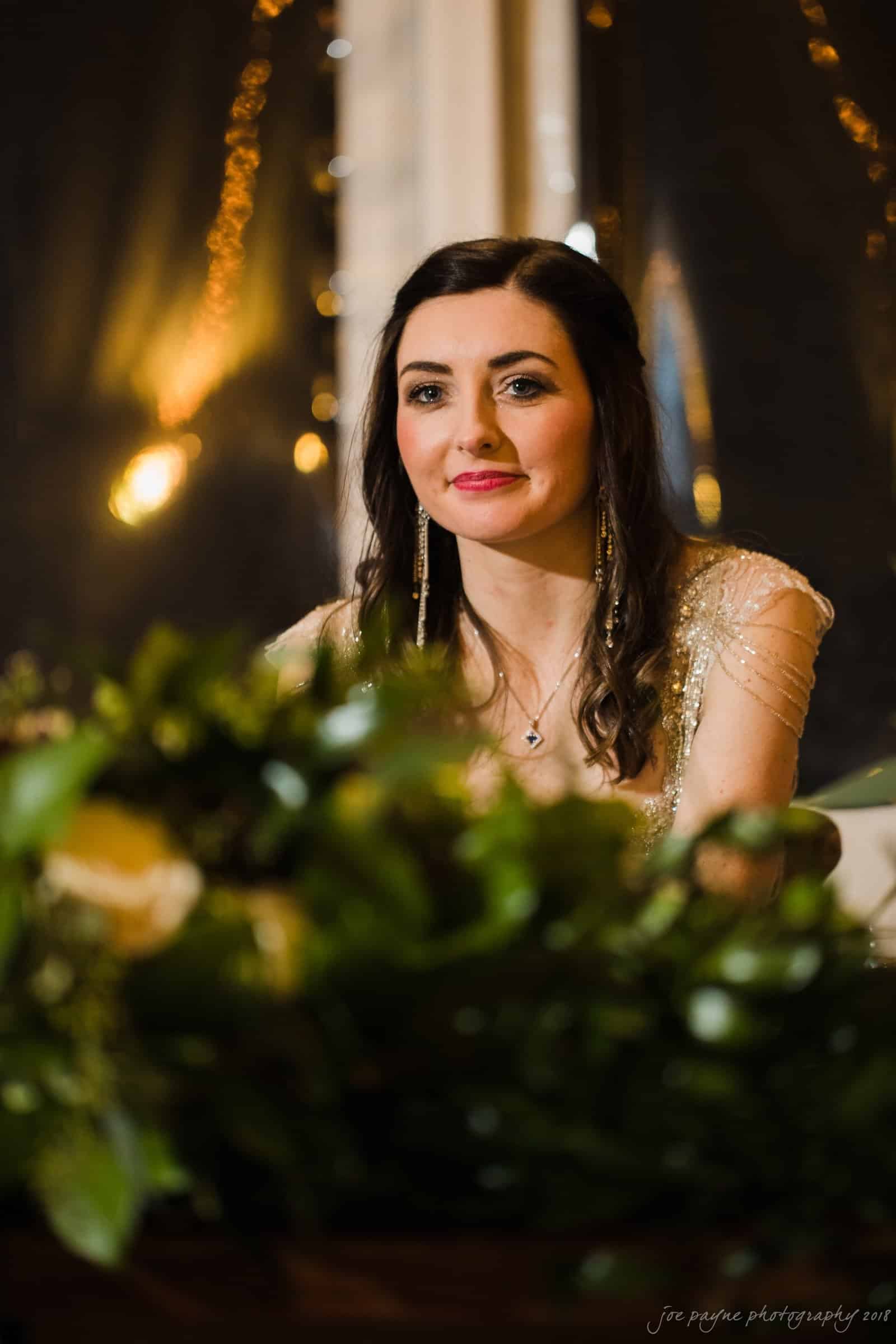 A woman with long dark hair and a gentle smile sits at a table adorned with green foliage. She wears a light-colored dress and long earrings. Soft, warm lighting creates a cozy atmosphere.