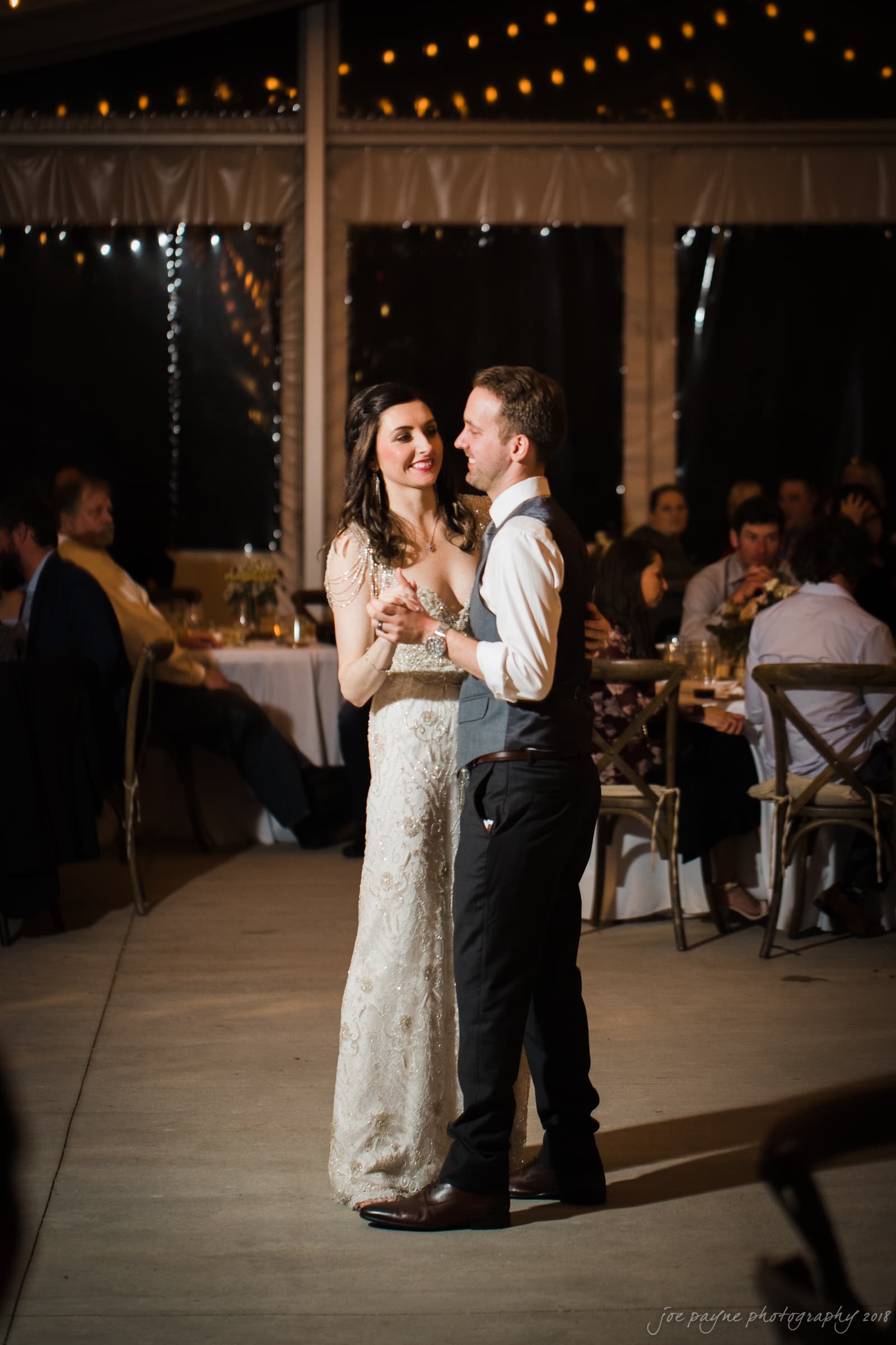 A bride and groom share their first dance at a wedding reception. The bride wears a lace gown, and the groom wears a vest and tie. Guests are seated at tables decorated with flowers and candles. String lights add a warm glow to the venue.