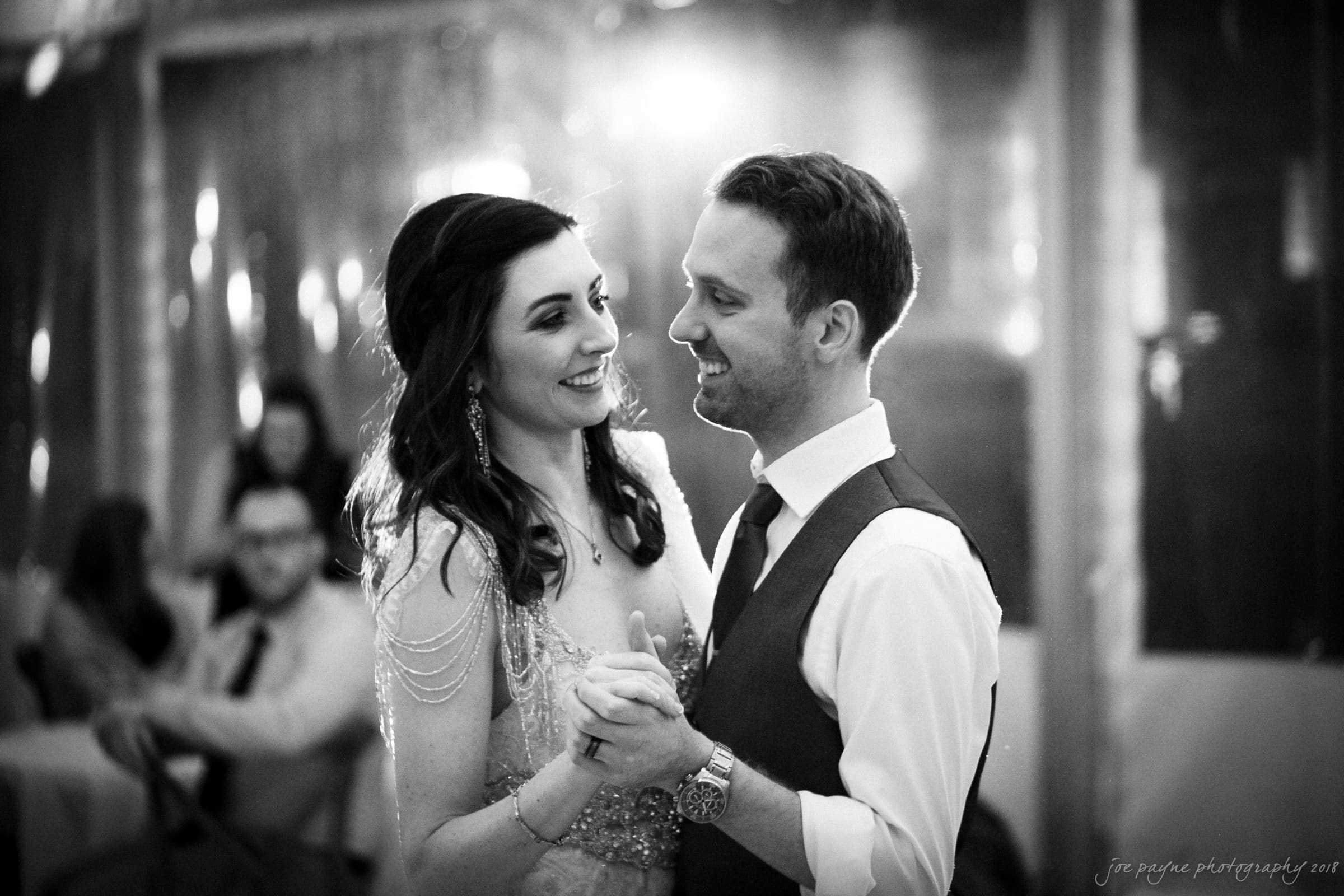 A couple is dancing closely at an indoor event. The woman is wearing a dress with decorative shoulder chains, and the man is in a shirt and vest. They are smiling at each other, with other guests blurred in the background. The scene is in black and white.