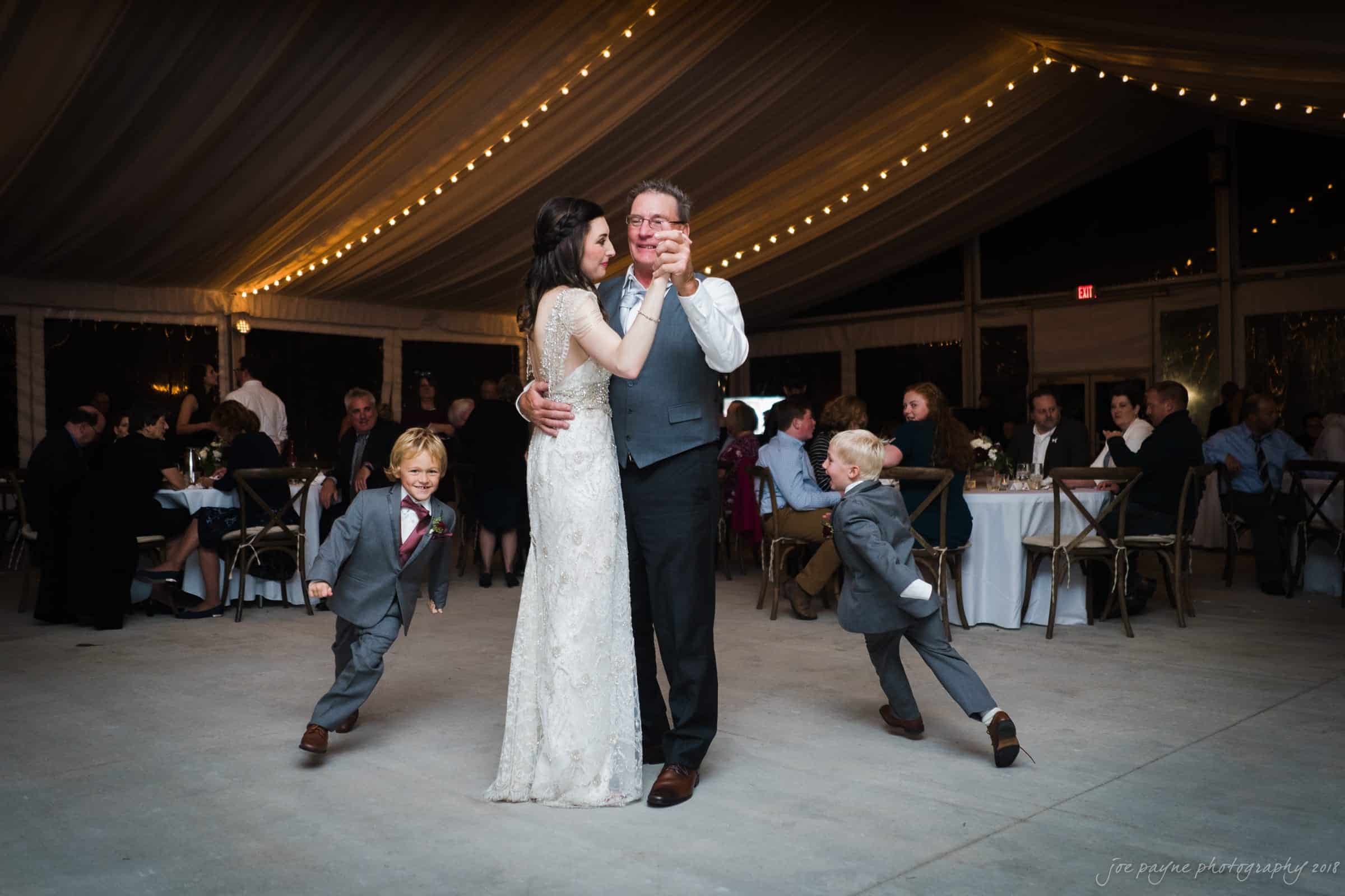 A bride and groom embrace on the dance floor under a tent with string lights. Two young boys in suits joyfully run around them. Guests are seated at tables in the background.