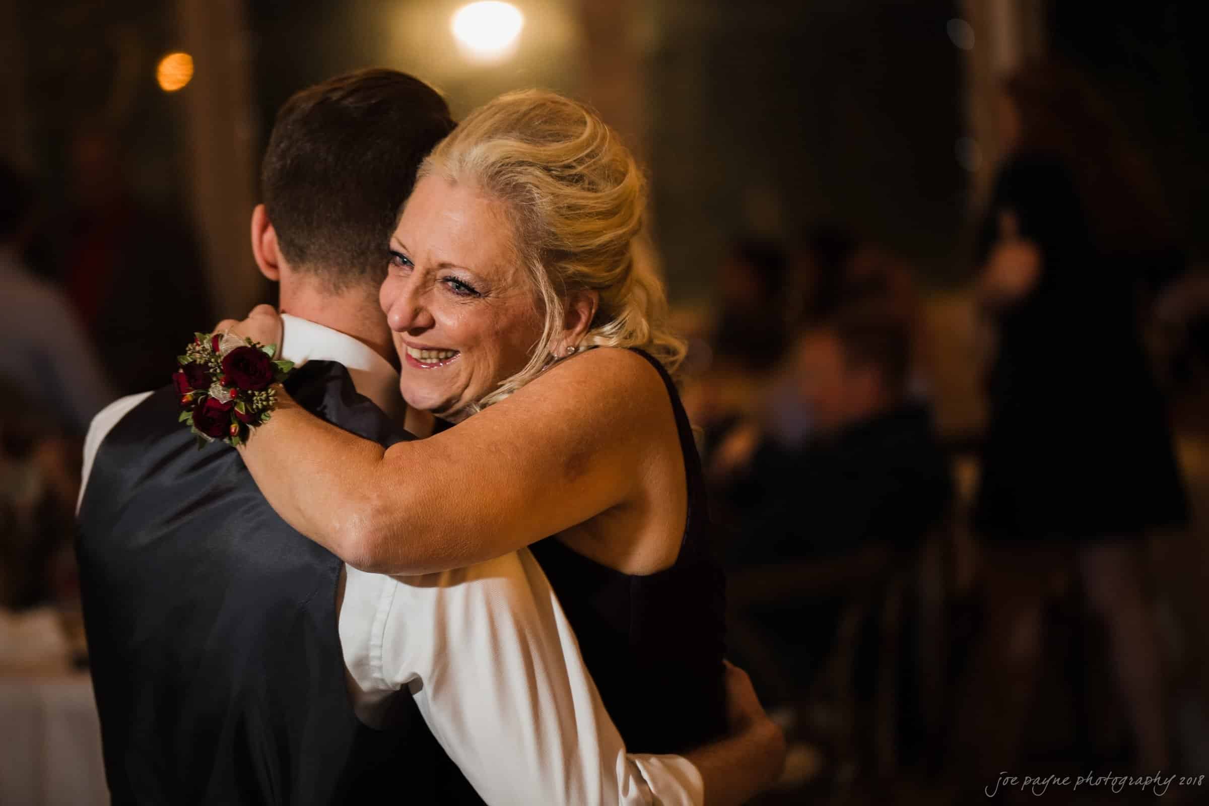 A woman with blonde hair smiles joyfully as she embraces a man in a formal suit on a dimly lit dance floor, surrounded by people. She wears a corsage on her wrist.