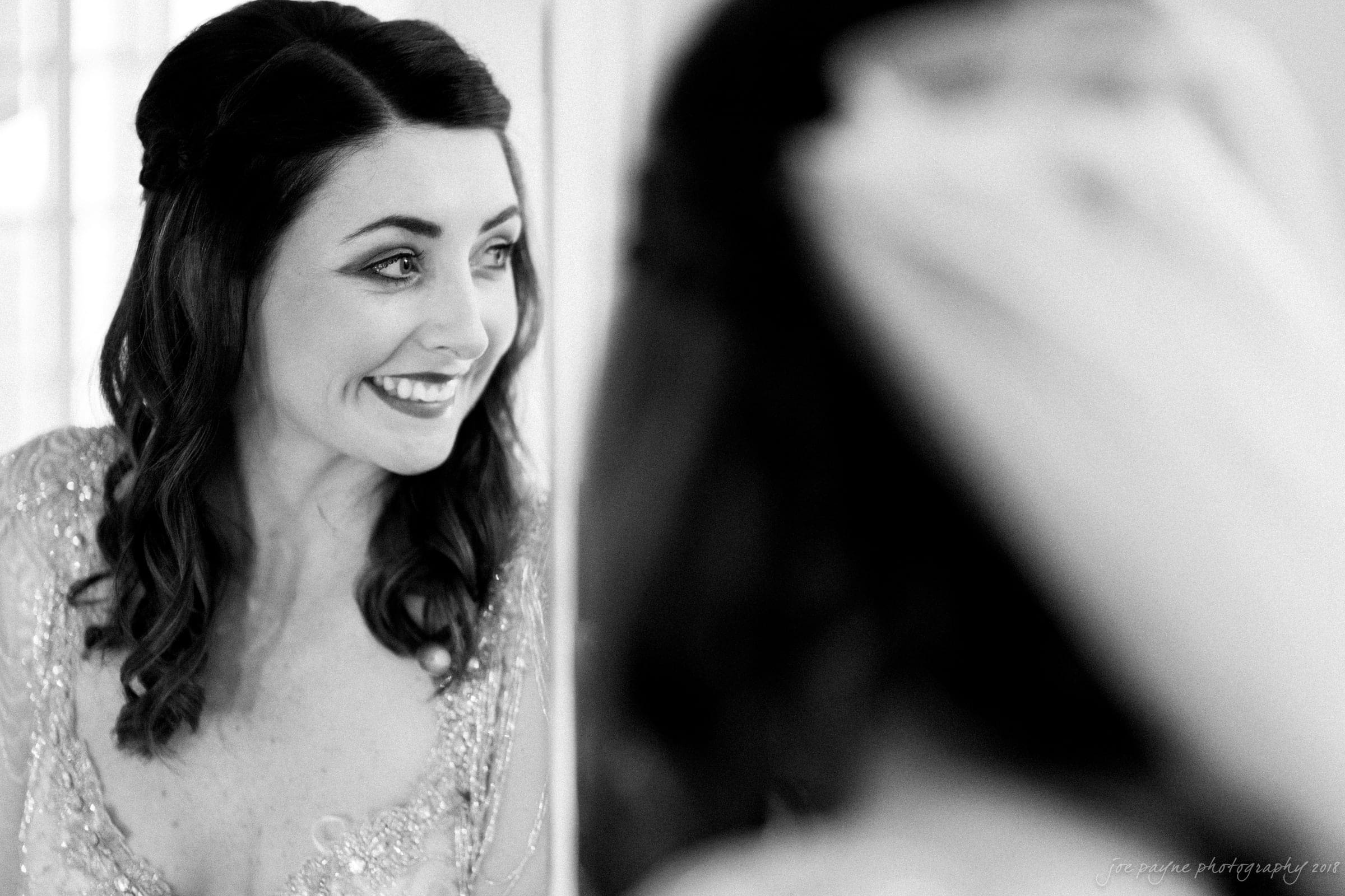 Black and white photo of a smiling woman looking at her reflection in a mirror. She has wavy hair and is wearing a dress with a beaded top. The image captures a joyful, candid moment.