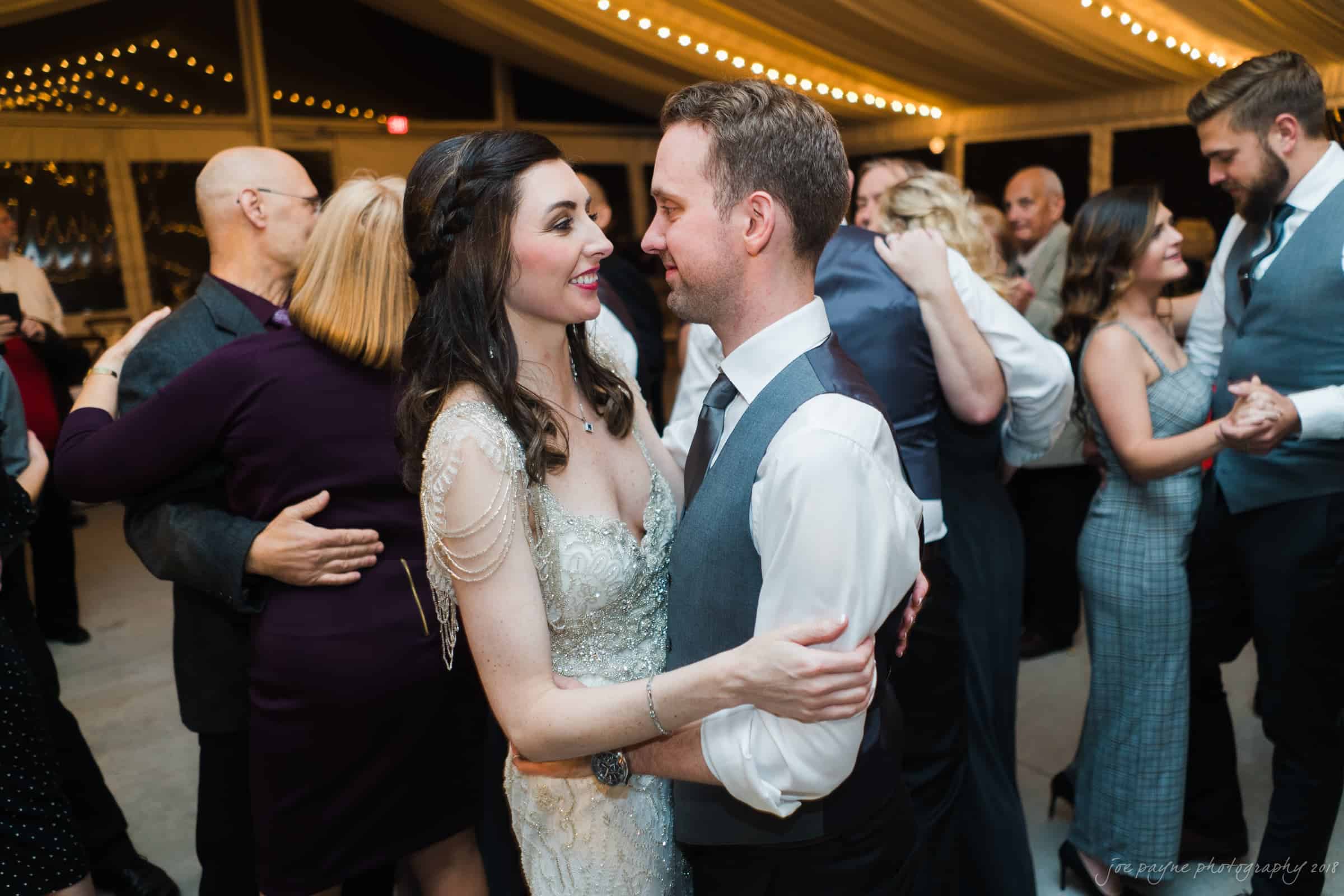 A couple dances closely, gazing into each others eyes, under a tent adorned with string lights. Other couples and individuals dance in the background, creating a lively, festive atmosphere.
