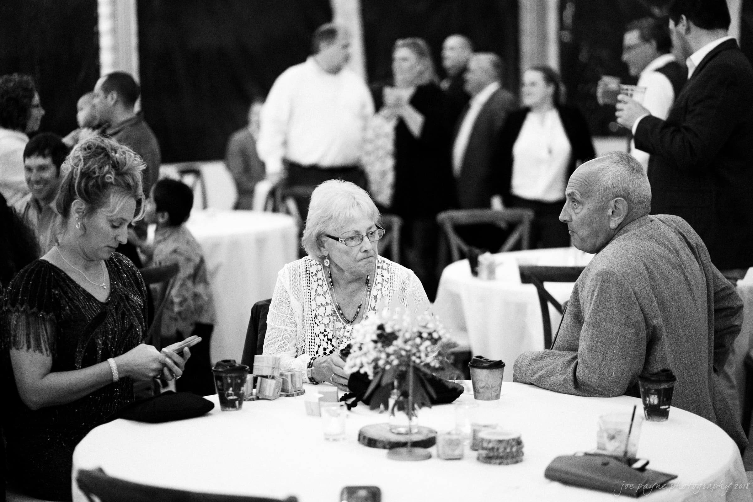A black-and-white photo of three people sitting at a round table during a social gathering. The woman on the left is using her phone, while the older woman and man beside her are engaged in conversation. Other guests and tables are visible in the background.