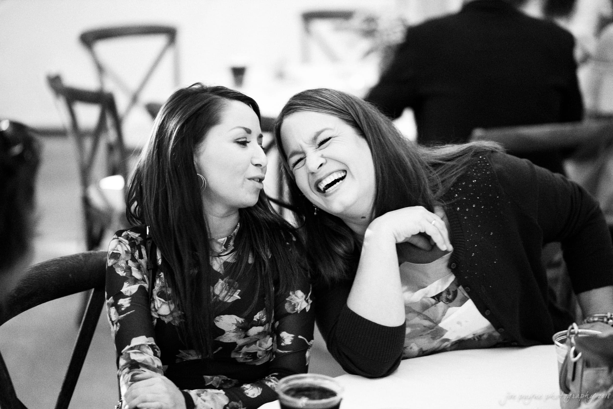 Two women seated at a table share a lighthearted moment. One is whispering while the other laughs joyfully. Both wear floral-patterned attire. The setting appears to be a social event, with blurred chairs and people in the background.