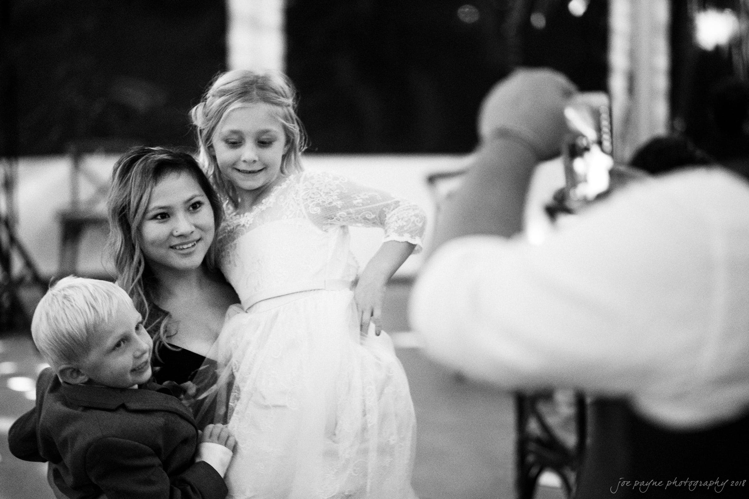 A woman smiles while posing with two children, a boy and a girl in a white dress. The boy is wearing a suit. They are being photographed by someone holding a smartphone. The setting is a warmly lit indoor space.