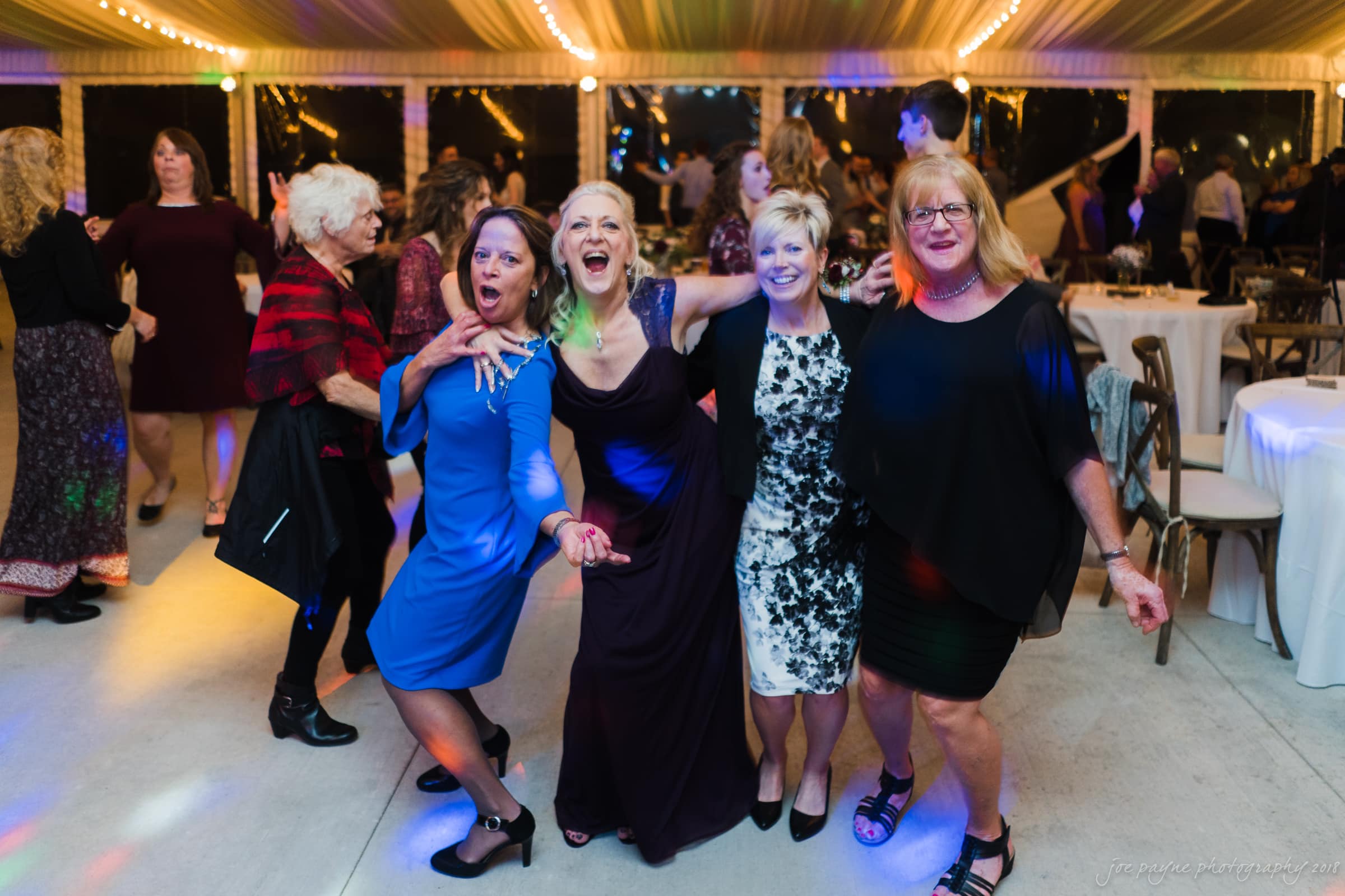 A group of four women, dressed in colorful evening attire, happily pose together on a dance floor. Behind them, other people are seen dancing in a festive, well-lit venue with tables in the background.