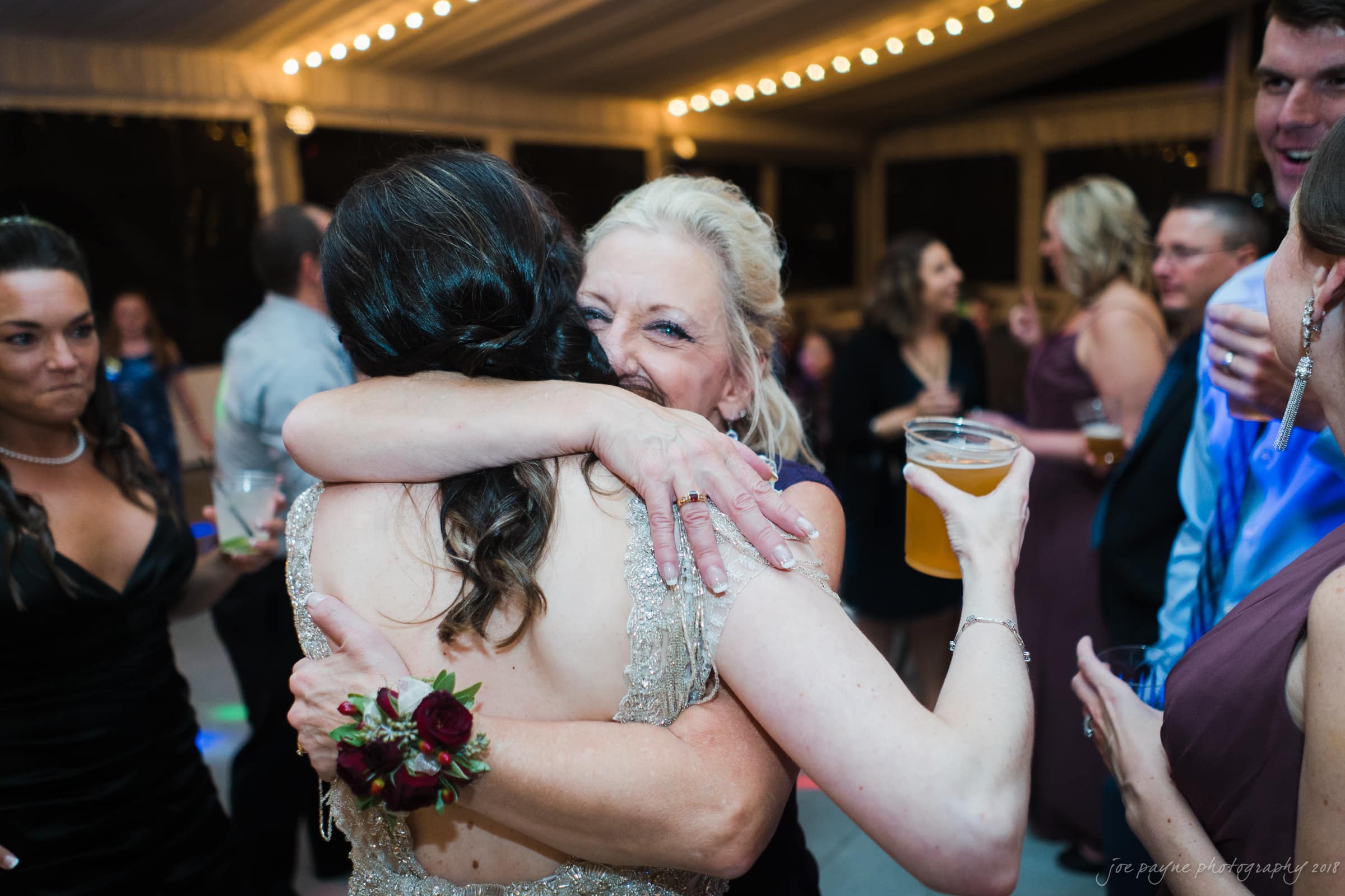 Two women are embracing at a lively event. One holds a drink while smiling happily. People in formal attire are mingling in the background, under string lights.