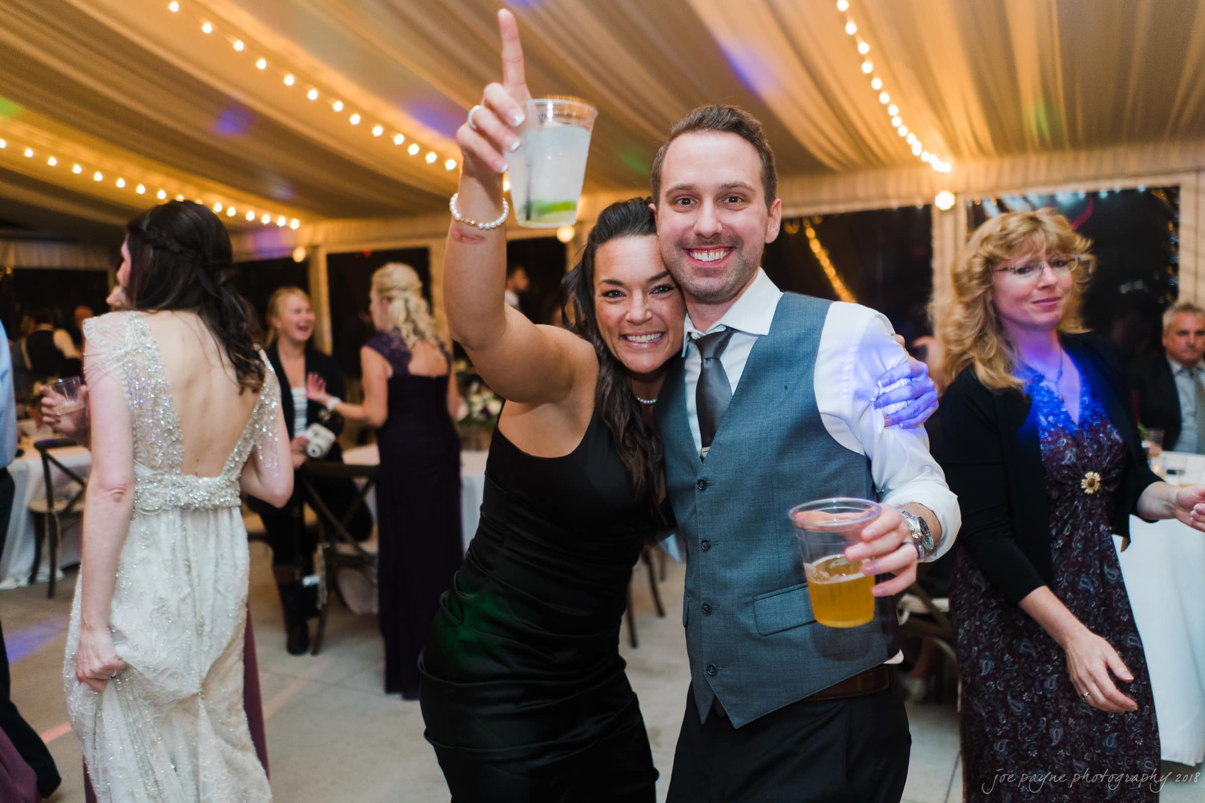 A joyful man and woman smiling and posing with drinks in hand at a festive indoor event. The room is warmly lit with string lights. Other guests are socializing in the background.