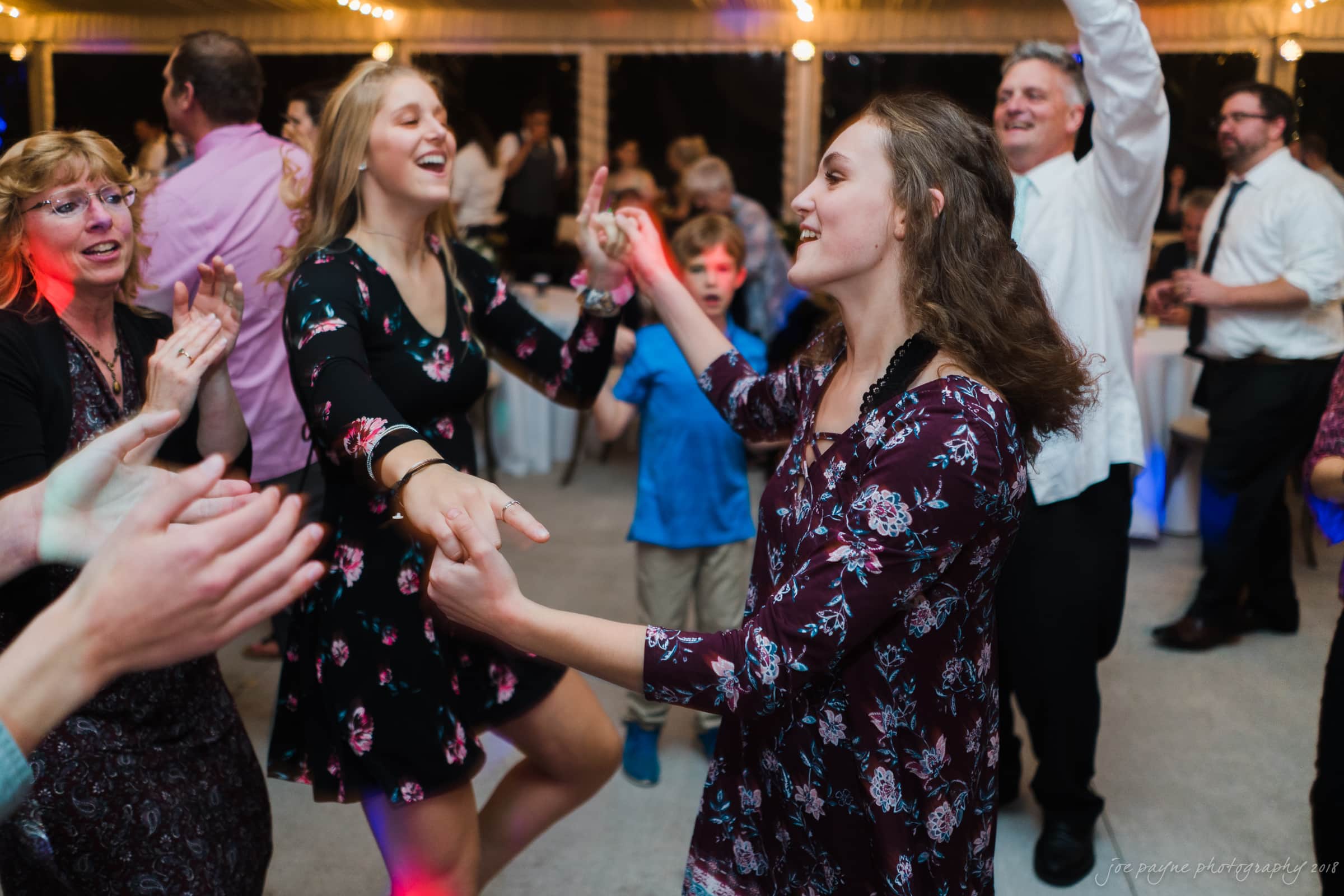 A lively scene at a dance event with people enjoying music. Two young women in floral dresses are dancing energetically in the center. A few adults and a child in the background are clapping and cheering under string lights.