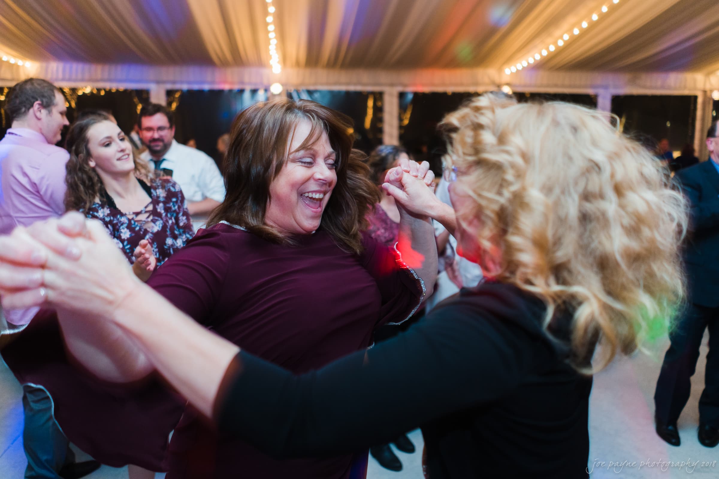 Two women joyfully dance together at an indoor event. They are surrounded by others, with warm lights illuminating the space. Both are smiling and appear to be enjoying the moment.