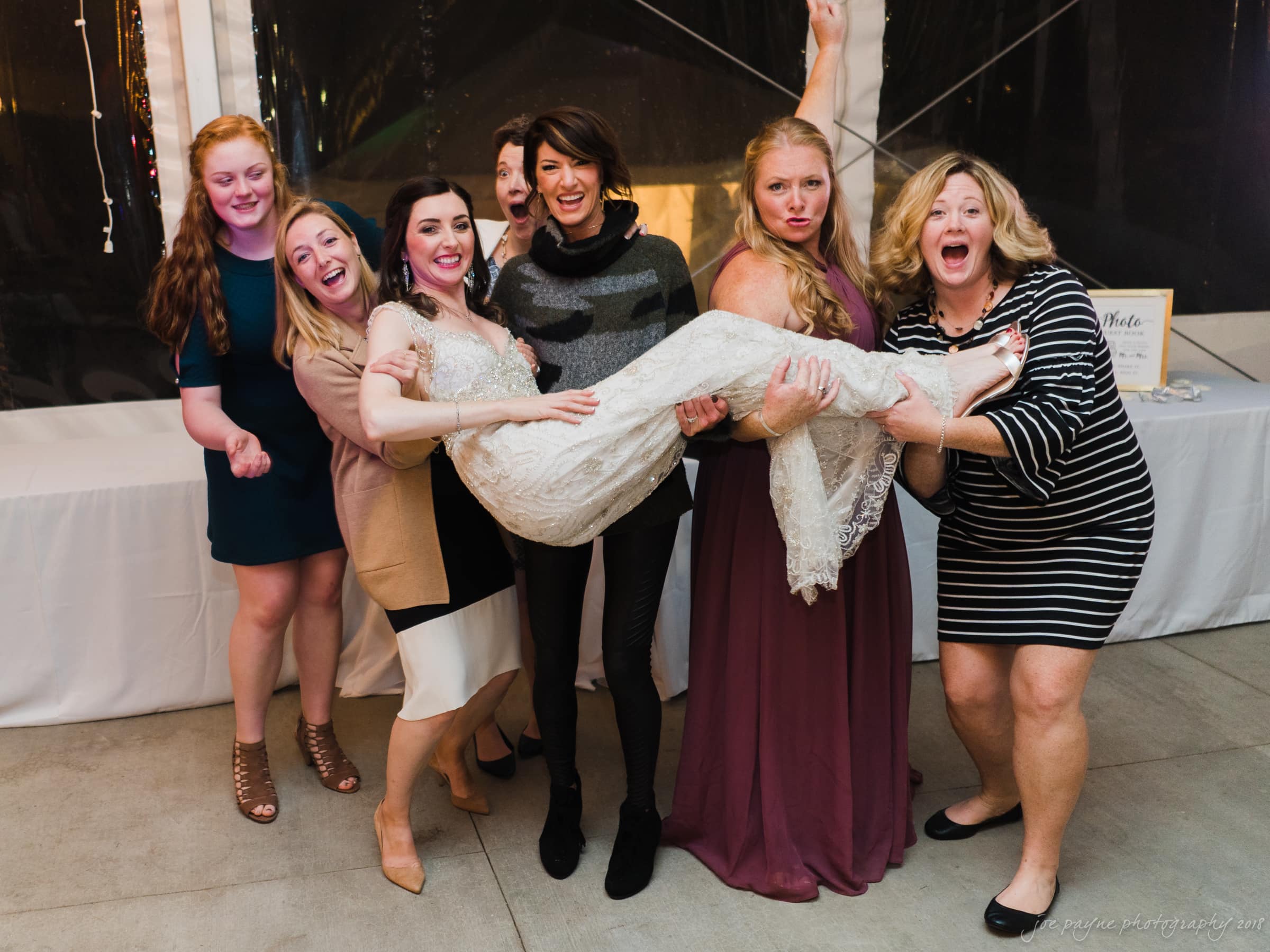 A group of six women is joyfully holding another woman in a white lace dress. They are indoors, and everyone is smiling and laughing. The atmosphere is celebratory and lighthearted.