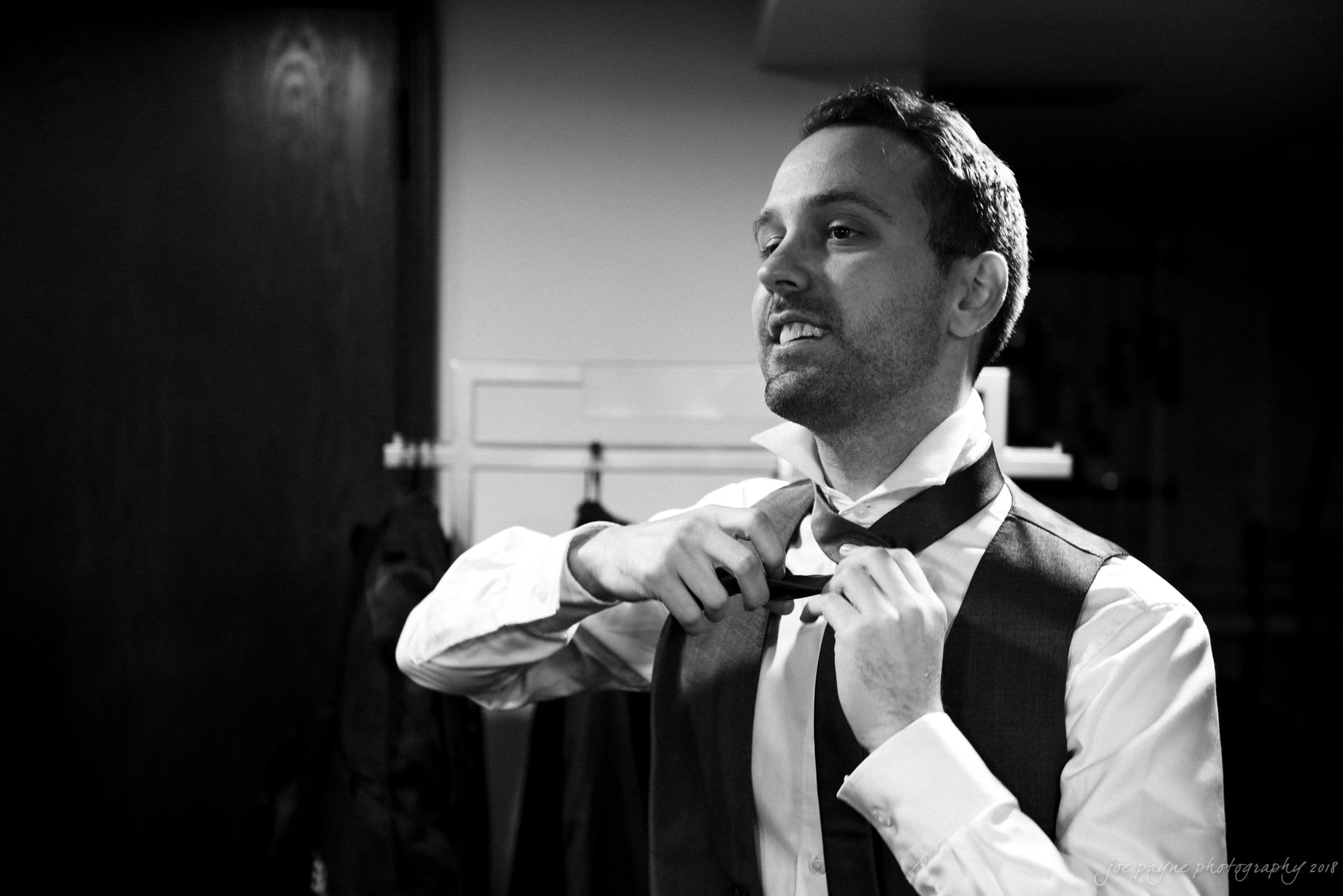 A man in a suit adjusts his tie in a dimly lit room, looking focused. A jacket hangs on a nearby rack. The black and white image gives a classic, timeless feel.