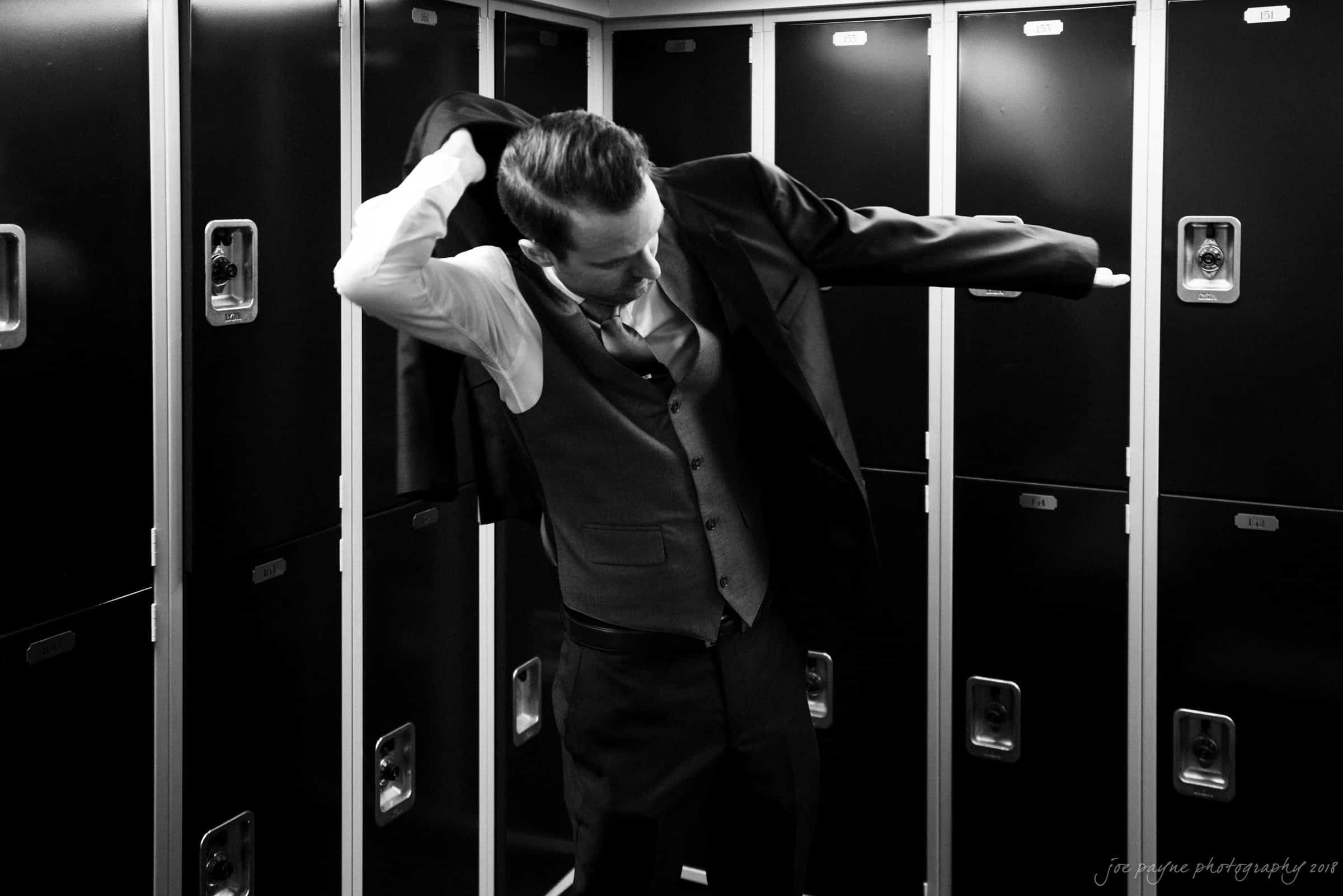 A person in a suit is putting on a jacket in front of a row of lockers. The image is in black and white, highlighting the contrast between the dark suit and the metal lockers.